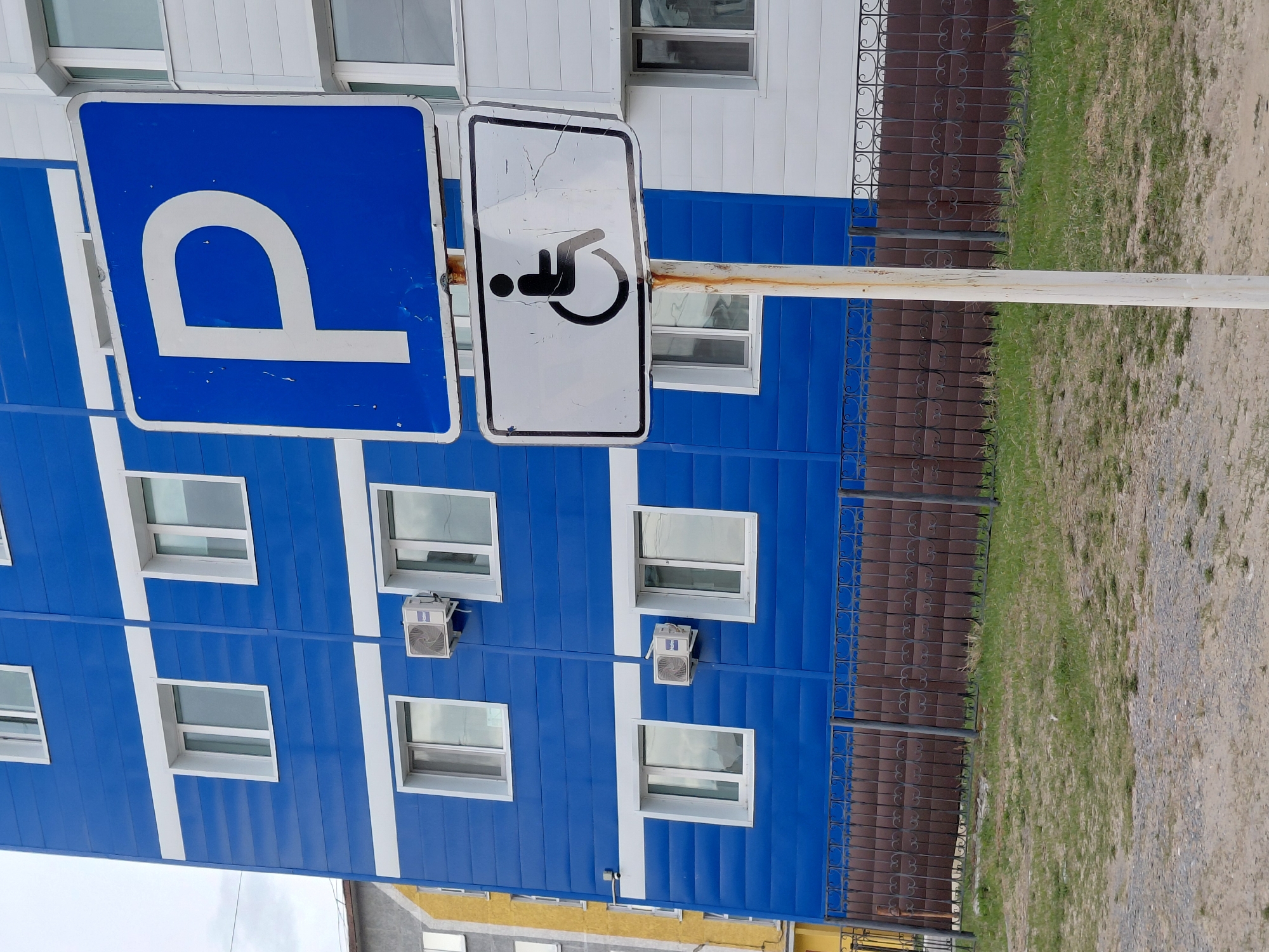 The image displays an outdoor scene, rotated 90 degrees clockwise from its natural orientation, showing a building, parking signs, and ground features.
In the foreground, a rusty metal pole supports two traffic signs, indicating a parking area, specifically for disabled persons. The left sign is blue with a large white 'P' (for Parking) and shows some wear and scuffs. The right sign is white with a black universal wheelchair symbol, also appearing aged with cracks and scratches on its surface.
Behind the signs stands a multi-story building, clad in blue siding with prominent white horizontal and vertical stripes, creating a grid-like pattern. Numerous rectangular windows with white frames are visible across the blue facade. Several white outdoor air conditioning units are mounted below some of the windows on the building. The very top of a different building, possibly white or light-colored, is visible above the blue building.
To the right of the pole, a dark, decorative metal fence with scrolled patterns runs parallel to the building. Beyond the fence, the ground is a mix of patchy green grass and bare dirt or gravel. The lighting suggests it is daytime, under a bright sky.
The scene depicts a designated parking area next to a building in what appears to be an urban or developed setting in Nadym, Russia.