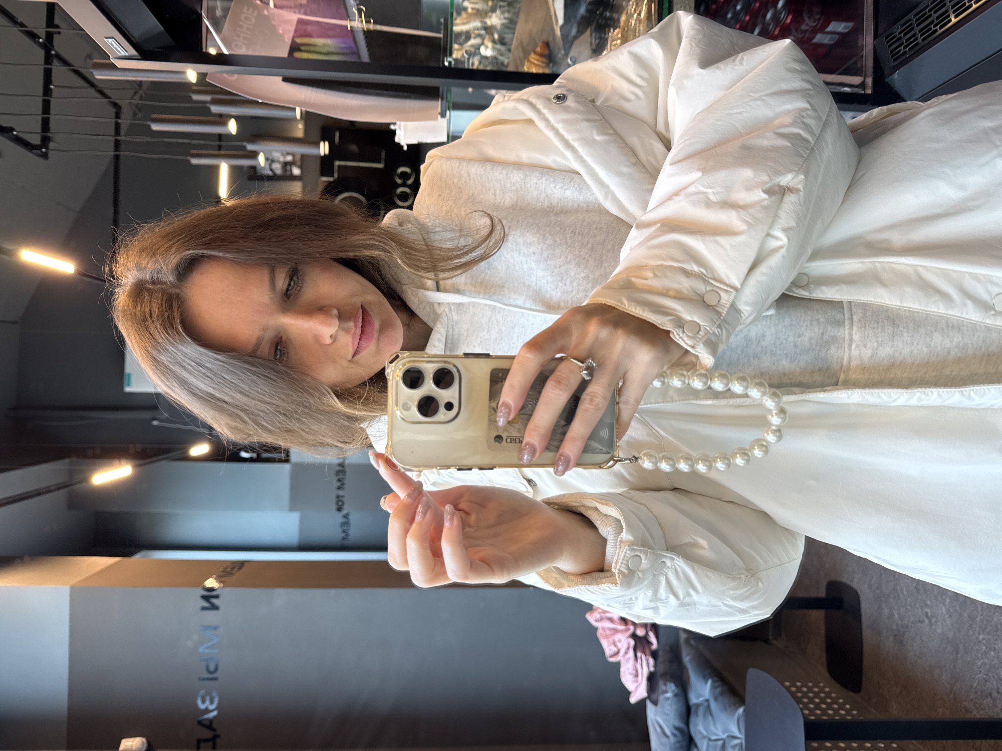 A woman is taking a selfie in a store in Moscow, Russia. She is holding a cell phone in her right hand, which has a string of pearls attached. Her left hand is extended in front of her. She is wearing a white puffy jacket over a light grey hooded sweatshirt, and a ring on her left hand. The reflection in the store window shows a grey wall with the name of the store written on it in Russian. Fluorescent lights are visible on the ceiling. A display case can be seen behind her.