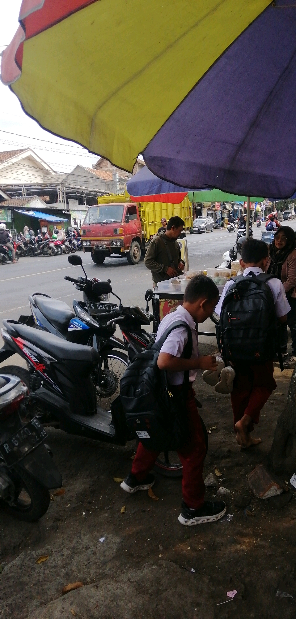 This image captures a street scene in Ajibarang Wetan, Indonesia, beneath a large, colorful awning. In the foreground, two young boys in school uniforms, one with a black backpack, walk away from the camera towards a food cart. One boy is carrying his shoes. Behind them, several motorbikes are parked. In the background, a red truck is stopped on the road, alongside other vehicles and a food stall. A man in a green jacket stands near the food cart, interacting with someone out of frame. Further in the background, buildings and more motorbikes indicate a bustling street environment. The ground is paved with concrete and scattered with leaves and debris.