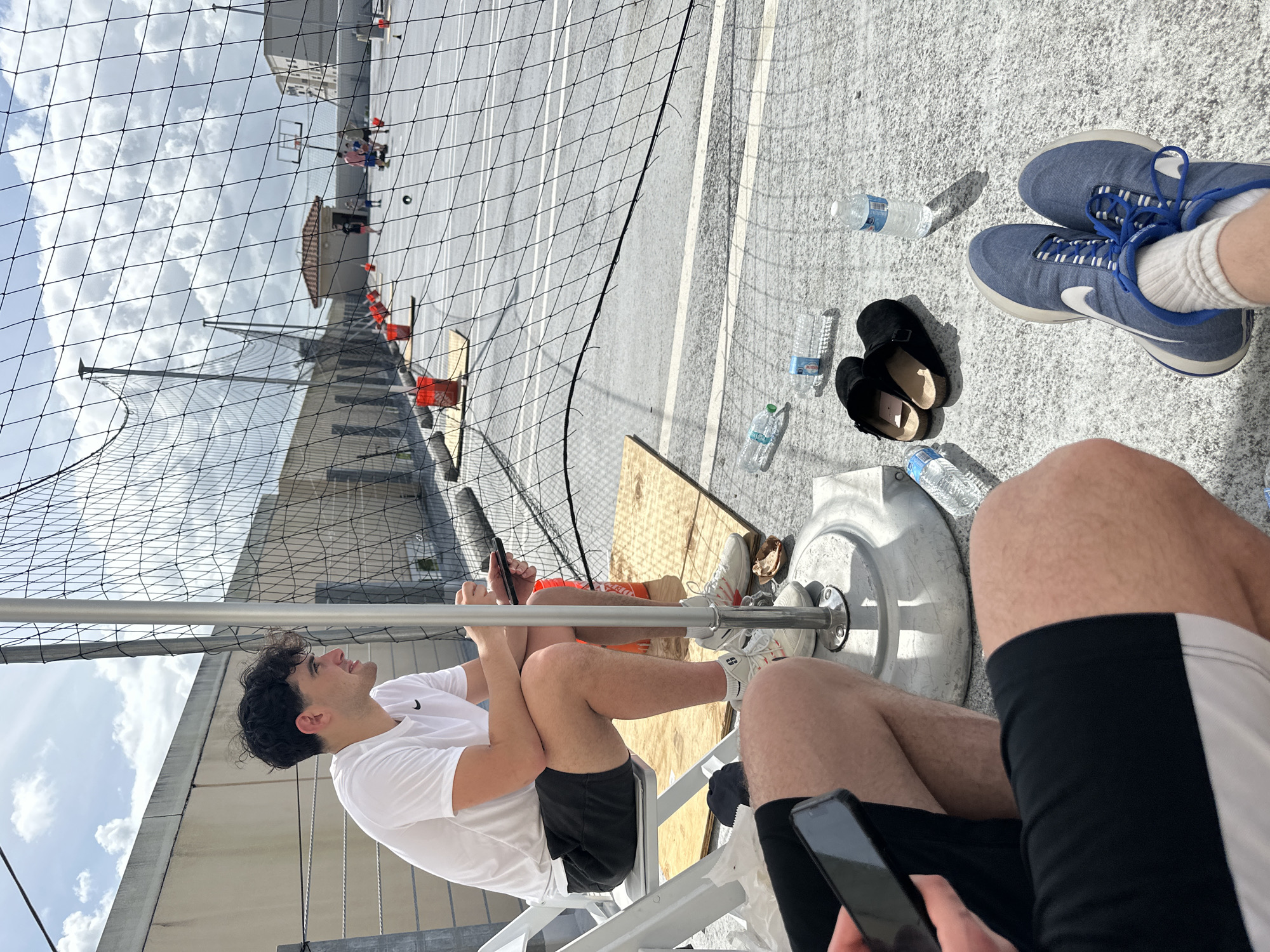 The image, taken in Aventura, United States of America, appears to show a recreational area, possibly a sports court viewed through a safety net. In the foreground, two people are seated. One is wearing a white t-shirt and black shorts and is holding a smartphone. The other, partly visible, is wearing black and white shorts and is also holding a smartphone. Nearby are two pairs of shoes (one blue and white trainers, one pair of black sandals), and several water bottles. In the background, through the net, a basketball court is visible with people engaging in play. Orange traffic cones are placed around the court. The surrounding area includes a building and the sky.