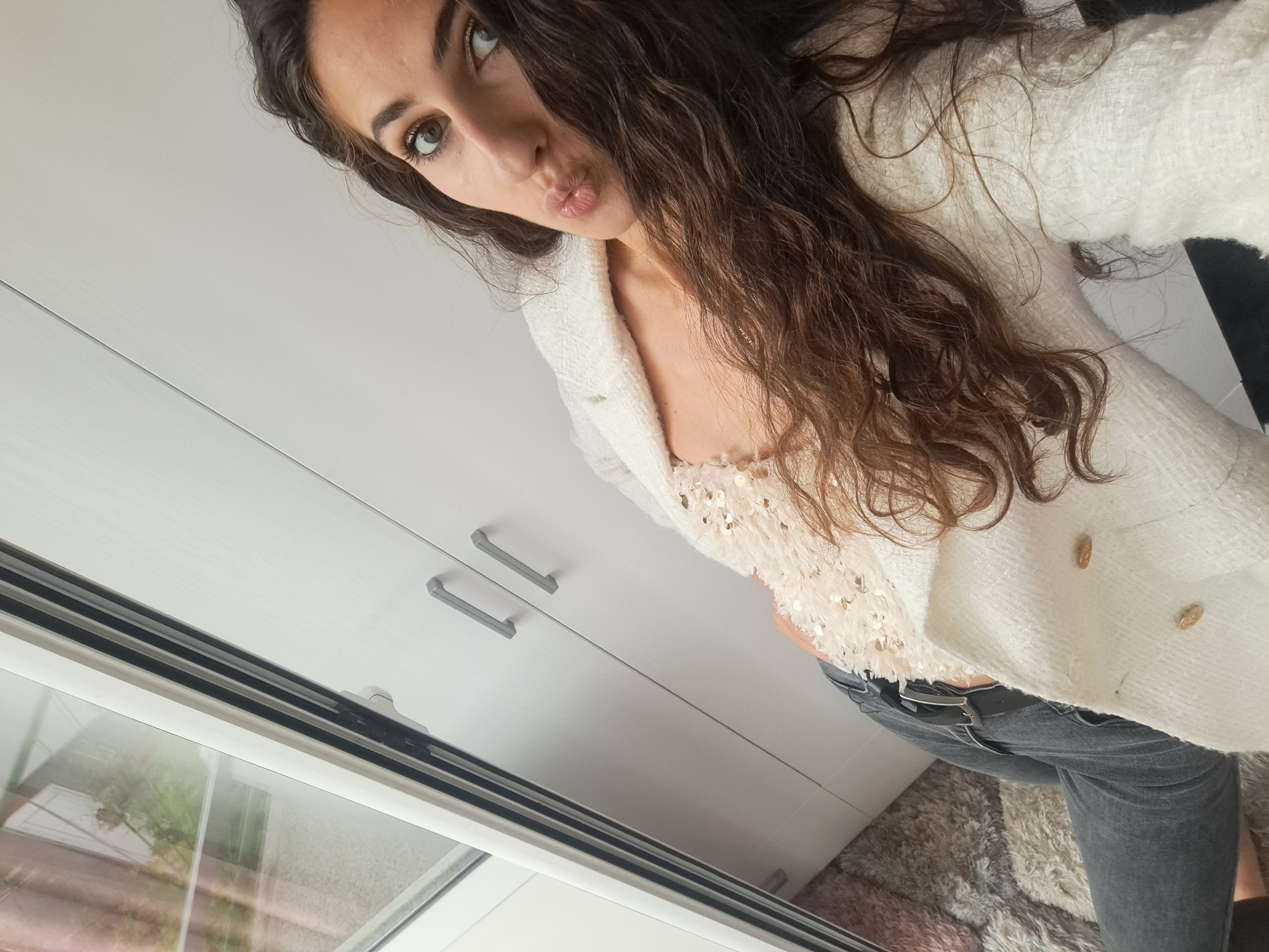 A woman with long, dark, curly hair, wearing a white textured jacket over a light-colored top with sequins and dark jeans, poses for a selfie indoors. She is leaning towards the camera, with her lips pursed. Behind her is a white cabinet with two grey handles and a window with reflections of greenery. A textured rug is visible on the floor. The scene appears to be in a domestic setting.