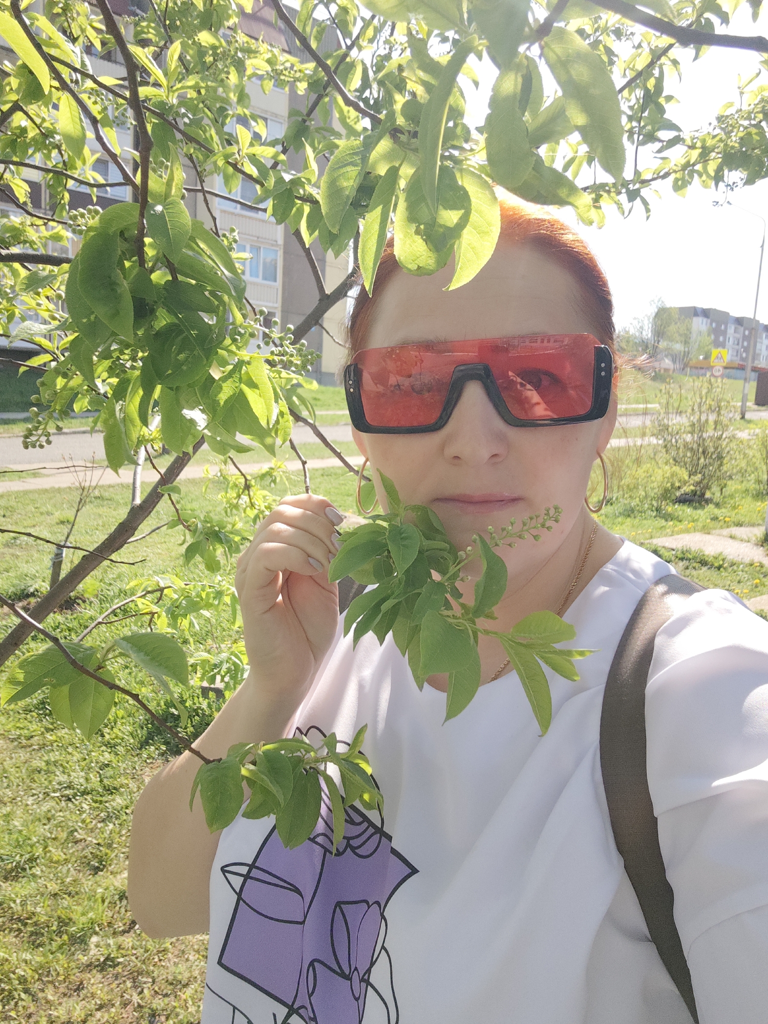 A woman with red hair stands outside in daylight holding a small leafy branch. She wears large, black-framed sunglasses with red lenses, gold hoop earrings, and a white T-shirt with a purple design. A gold necklace is visible around her neck. A dark-colored backpack strap is over her right shoulder. The background includes a green lawn, a tree with bright green leaves, and a multi-story building in the distance, indicating an urban environment. The location is Марковский, Russia, as indicated by the information provided with the prompt.