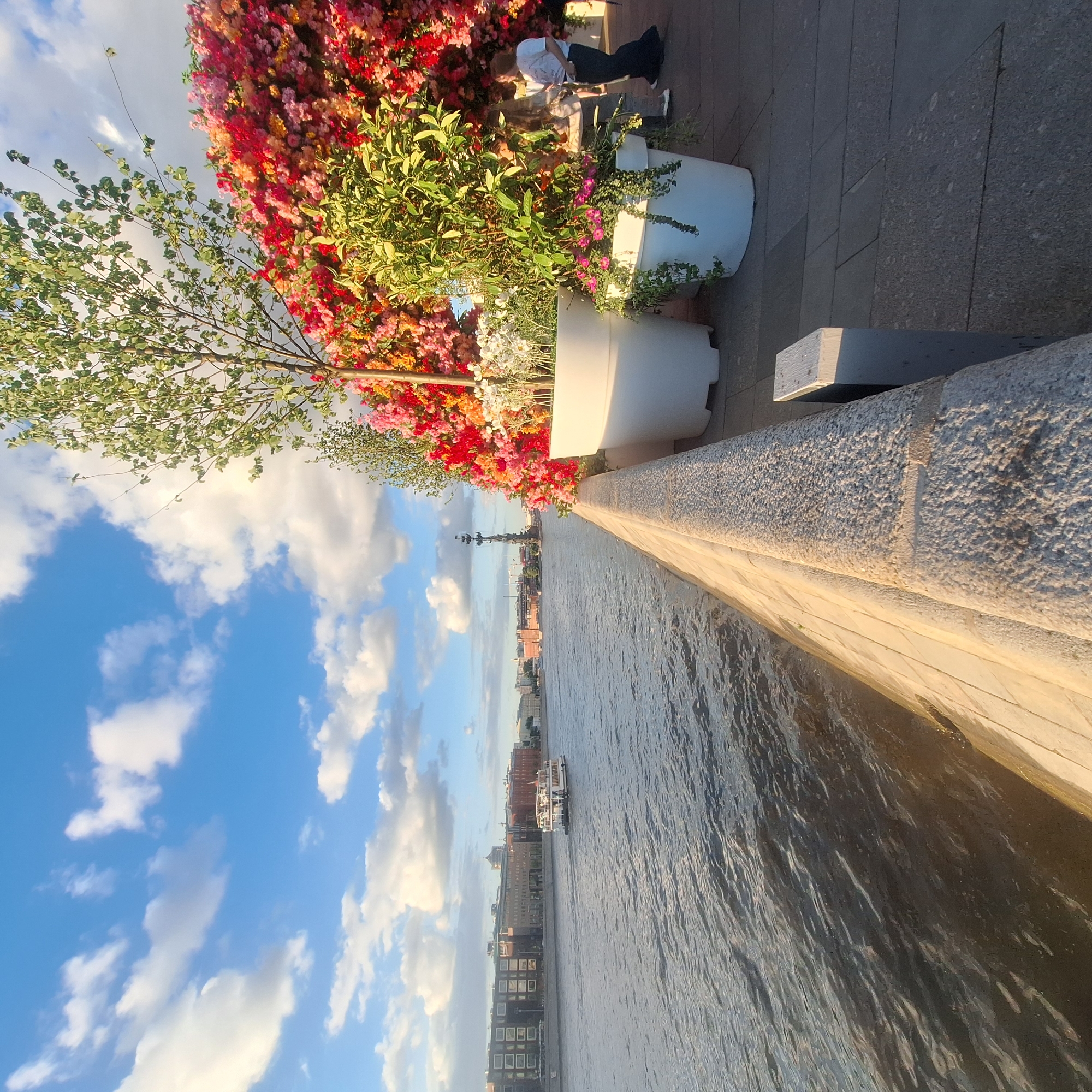A sideways medium shot shows Moscow, Russia, on a partly cloudy day, with a river, city buildings and flowers in view. In the foreground is a river, with a boat in the middle ground. Tall buildings line the shore and the sky is visible. Along the top of the frame is a stone wall with a cement bench in front. White flower pots with green and colorful flowers line the top of the wall. People are sitting and standing near the flowers.