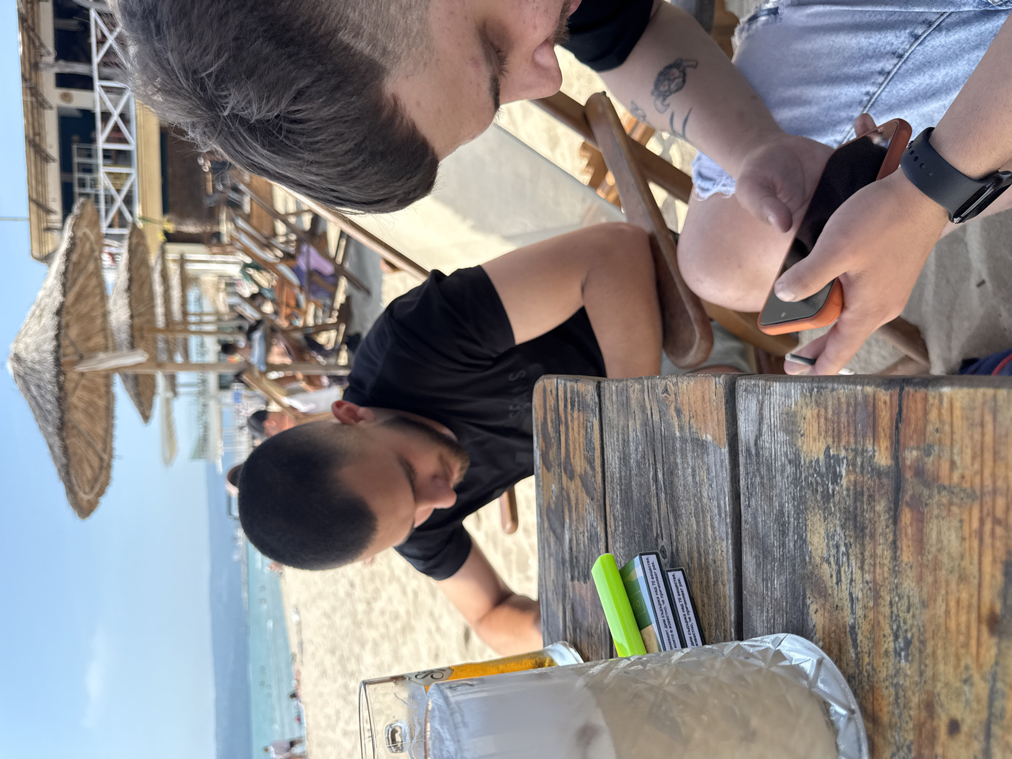 The image captures an outdoor scene at a beachside establishment in Varna, Bulgaria, under bright daylight with a clear blue sky. Two men are seated at a rustic wooden plank table in the foreground.
The man on the left, with dark, short-cropped hair and a beard, wears a black t-shirt and is seen leaning forward, looking down towards the table. To his left, a tall, textured glass filled with a light, frothy beverage, possibly an iced drink, is prominently displayed. Behind it, a beer glass, or stein, containing a golden-yellow liquid, is partially visible.
In the center of the table, two packs of cigarettes are stacked. The top pack displays Cyrillic text, reading: "ОНЕВИЯТ ДИМ СЪДЪРЖА НАД 70 ВЕЩЕСТВА, КОИТО Е ИЗВЕСТНО, ЧЕ ПРИЧИНЯВАТ РАК." (Translation: "THE SMOKE CONTAINS OVER 70 SUBSTANCES WHICH ARE KNOWN TO CAUSE CANCER."). A bright neon-green cylindrical object, possibly a vape pen or lighter, rests on top of the cigarette packs.
The man on the right, partially visible, has dark, short-cropped hair. His right arm, adorned with a dark tattoo of a turtle-like figure, is holding an orange-cased smartphone horizontally with both hands, indicating active use. He wears light-colored, possibly ripped denim shorts and a black smartwatch on his left wrist. He is looking down intently at his phone. Both men are seated in wooden beach chairs.
The background reveals a sandy beach with numerous individuals, some walking, others relaxing on the sand. The turquoise sea extends to the horizon, meeting a distant, hazy coastline. Several straw-thatched umbrellas are scattered across the beach. Further back, a two-story building with a white railing and a blue section is visible, suggesting a beach bar or restaurant.