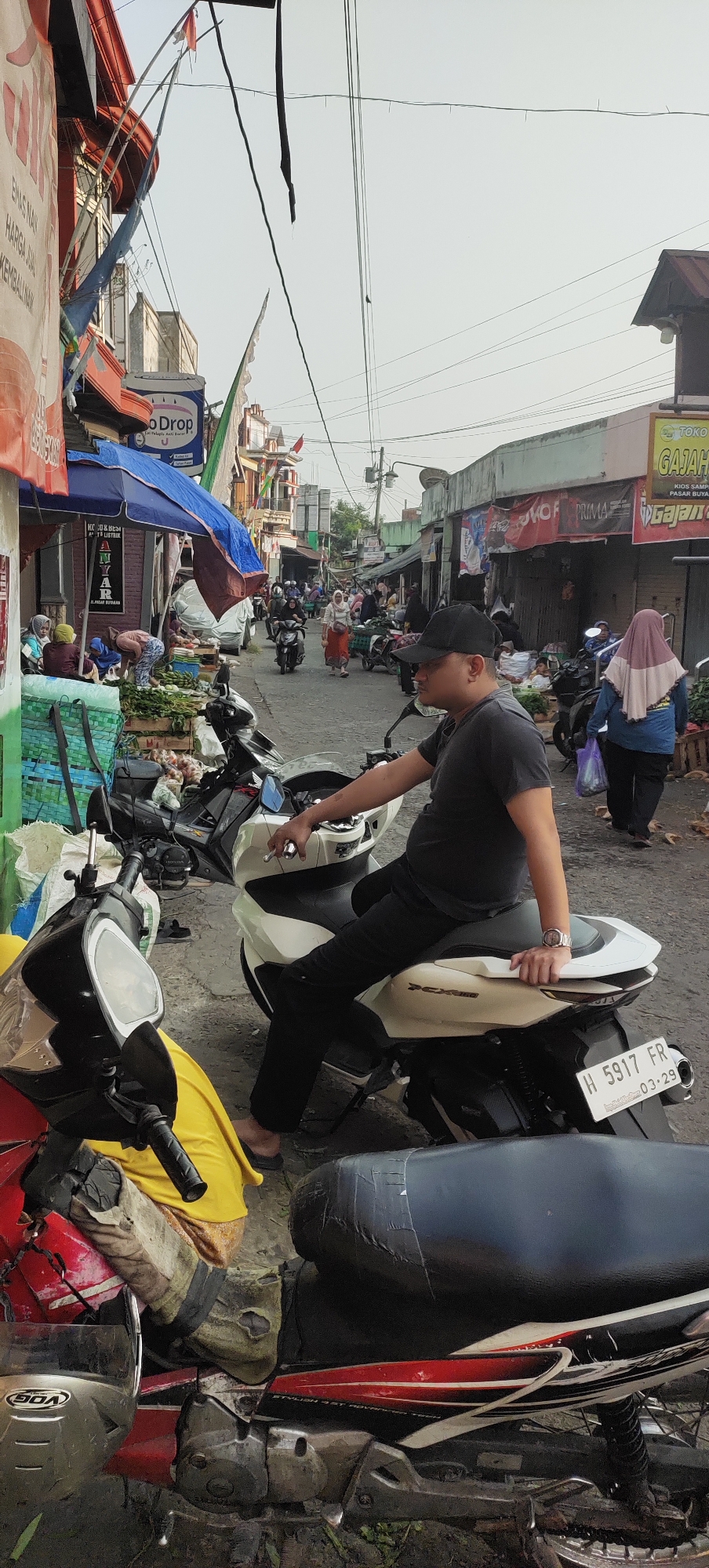 The image depicts a bustling street scene in Karangtowo, Indonesia, likely a market or commercial area, with people and motorcycles. In the foreground, a man wearing a black t-shirt and a black baseball cap is seated on a white scooter. He is looking towards his left, with his left hand resting on the handlebars and his right hand appearing to be near his mouth, possibly holding a cigarette. The scooter has a license plate reading "H 5917 FR 03-29". To the left of this scooter, another motorcycle, predominantly red, is partially visible, with a silver helmet resting on its seat.
The background shows a street lined with shops and stalls. Several people are walking or riding motorcycles along the street. Some individuals are seen wearing headscarves. Stalls display various goods, including what appear to be fruits and vegetables. Signs with text are visible on some buildings and stalls, including "TOKO GAJAH" which indicates a shop name. Overhead wires crisscross the sky, common in urban environments. The overall atmosphere suggests a lively daytime activity in a local Indonesian setting.