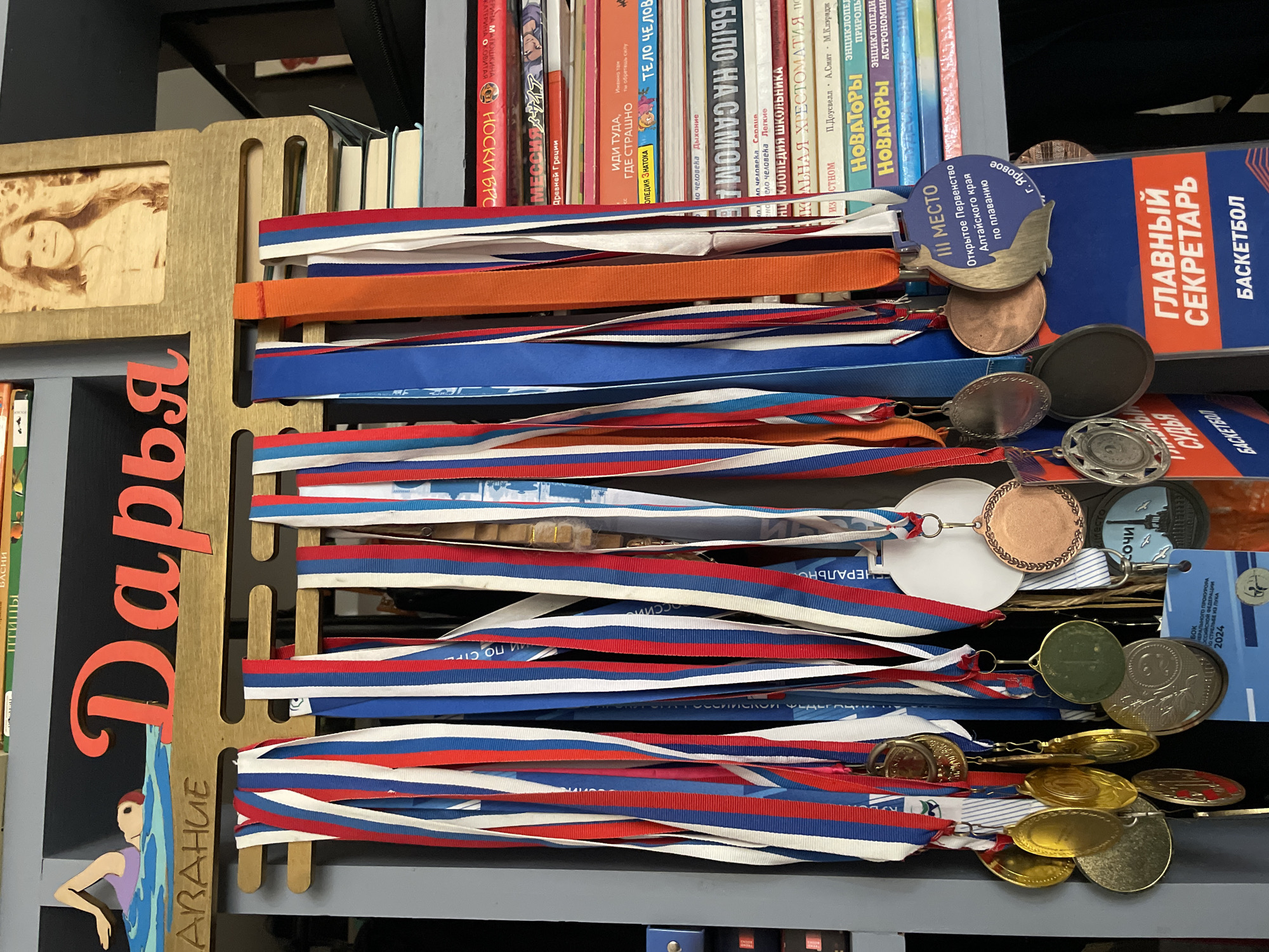 The image shows a shelf filled with books and medals. The books are of various sizes and colors, with titles in Russian, including textbooks and literature. The medals are hanging from ribbons in the colors of the Russian flag (white, blue, red), and some are gold, silver, or bronze. There are several medals from Сочи(Sochi, Russia). One medal and a book indicate the subject of basketball. There is a wooden frame with the name "Daria" written on it, along with a picture of a swimmer.