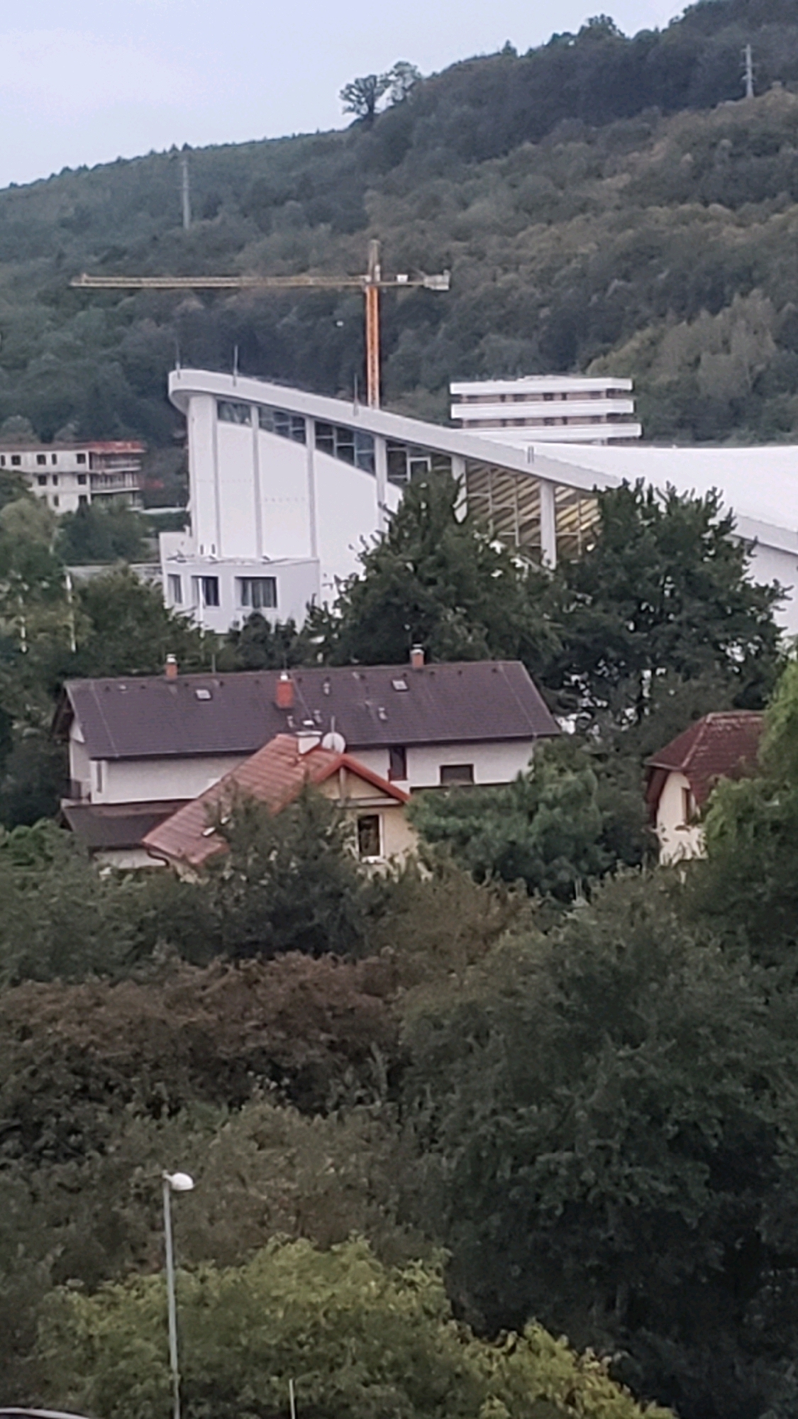 The image captures a residential area with a large modern building and a construction crane in the background, set against a backdrop of a tree-covered hillside. In the foreground, two houses with dark tiled roofs are visible, partially obscured by lush green trees. One of the houses has a lighter colored facade, while the other appears to be a lighter color with red tiled roof extensions. The larger building in the midground features a prominent white, curved roofline with visible windows. A yellow construction crane stands tall on the wooded hillside behind this building. The scene is likely located in Prešov, Slovakia, a city known for its historical architecture and surrounding natural landscapes. The overall impression is one of a developing urban area, blending modern construction with residential housing and natural elements.