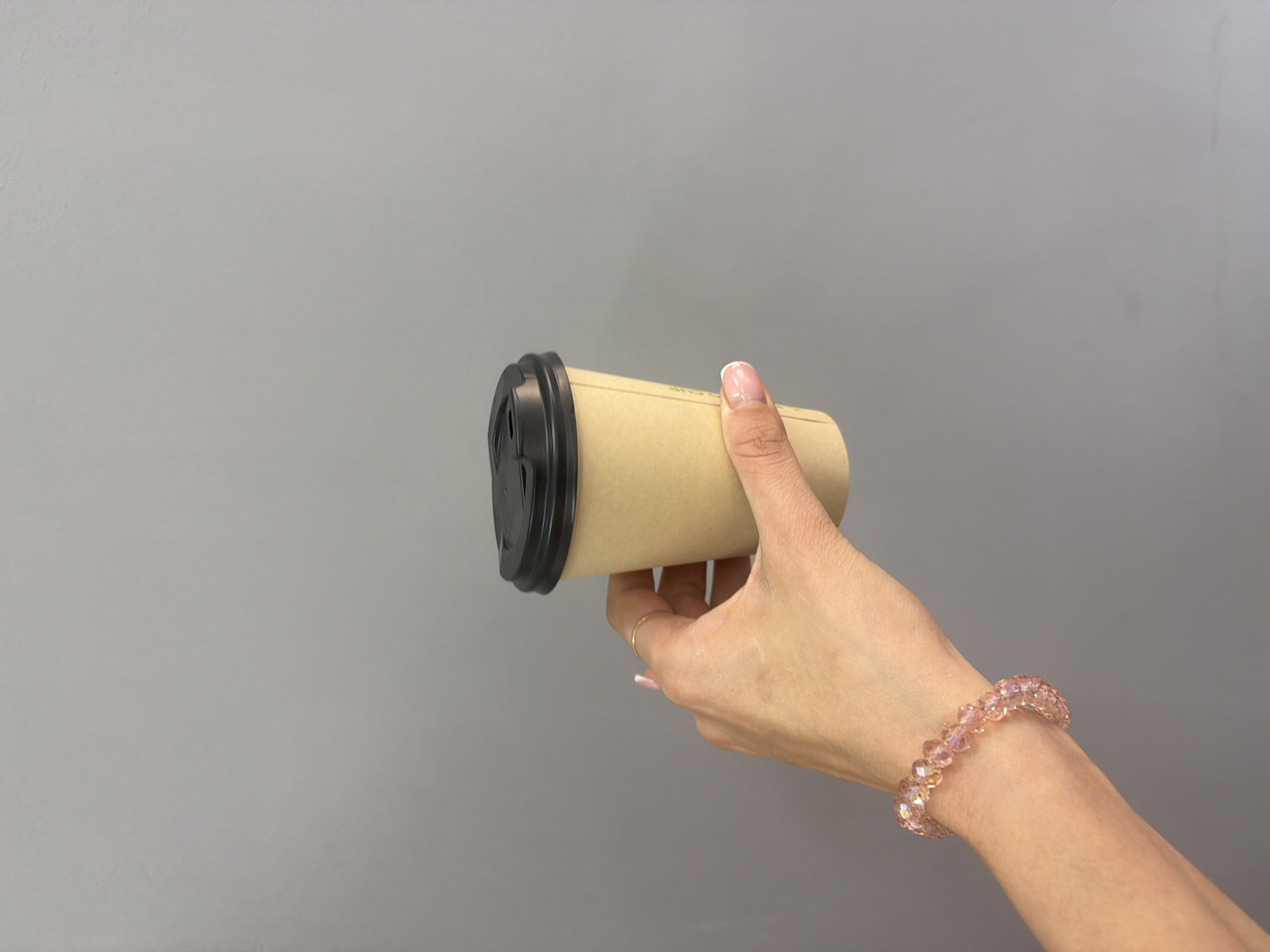 In Kyiv, Ukraine, a woman's hand wearing a pink crystal bracelet and a thin gold ring is holding a brown paper coffee cup with a black lid against a plain grey wall. The cup has some faint green text on it. The woman's fingernails are manicured with a French tip. The image is a close-up shot, focusing on the hand and the coffee cup.