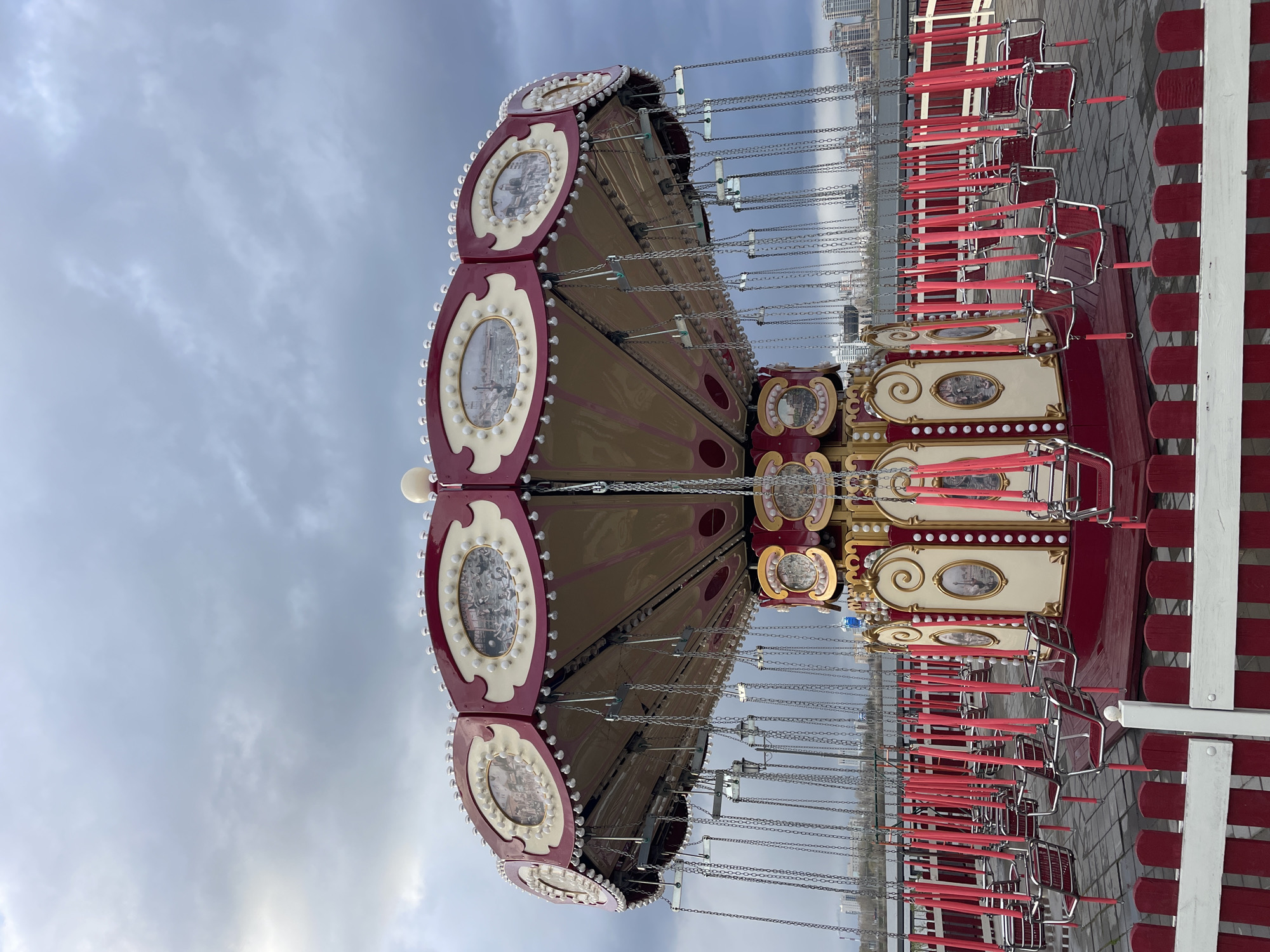 The image captures a detailed view of a large, ornate swing carousel, also known as a chair swing ride, under an overcast sky in Kazan, Russia. The ride is stationary, with no people visible on or around it, suggesting it is either closed, awaiting operation, or between rides.
The central structure of the carousel features a multi-tiered, rotating platform adorned with decorative panels. These panels are primarily deep maroon/red and cream/off-white, intricately designed with oval and circular insets. Each inset contains a small, detailed picture, possibly depicting historical scenes or landscapes, and is framed by numerous small, round, white lights or bulbs. Golden accents and scrollwork further enhance the ride's classic fairground aesthetic.
Numerous individual swings, suspended by multiple silver chains, hang from the main structure. Each swing has a bright red seat, often with a mesh pattern, supported by a metal frame. The swings hang vertically, indicating the ride is not in motion.
The setting shows a cloudy, light grey sky covering the upper portion of the image. The ground beneath the ride is paved, possibly with patterned bricks or cobblestones. In the distant background, a blurry cityscape with tall buildings is visible through and above the chains, providing a subtle urban context. The overall scene is quiet, showcasing the colorful and decorative amusement ride against the subdued weather. No discernible text is visible on the ride or in the surroundings.