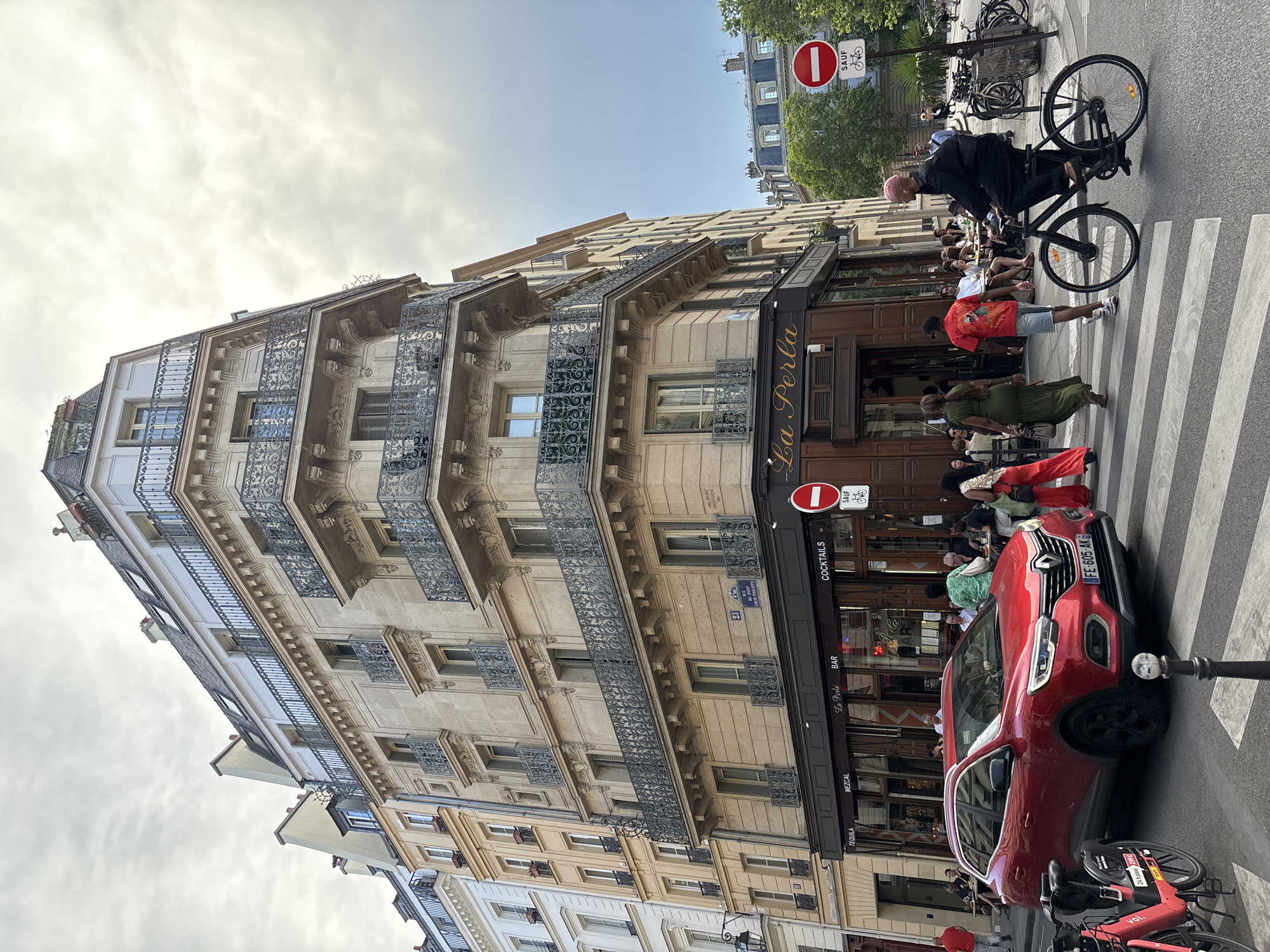 The image shows a street scene in Paris, France, likely in the area of a restaurant or bar called "La Perla." A red Renault SUV is stopped at a crosswalk, indicating traffic is present. Several people are visible on the sidewalk and entering or exiting the establishment. A person on a bicycle is also crossing the street. The architecture features typical Parisian buildings with wrought-iron balconies and ornate facades. A traffic sign with a red circle and a horizontal bar indicates a prohibition, possibly for vehicles. The sky is overcast, suggesting a cloudy day. The overall scene depicts a bustling urban environment in Paris.