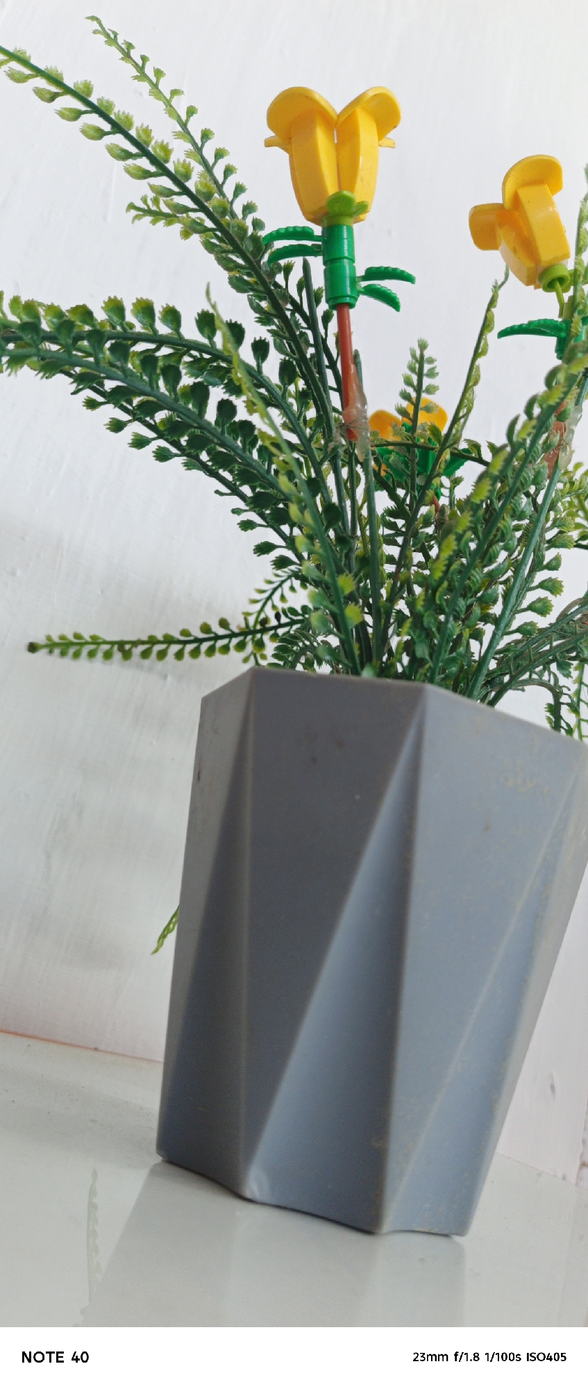 A close-up shot depicts a gray, geometric pot containing artificial plants with yellow flowers. The pot sits on a white surface, likely a table or shelf, against a textured white wall. The lighting is bright and appears to be natural. The photo's metadata, "NOTE 40 23mm f/1.8 1/100s ISO405," is visible at the bottom. The image does not include people, actions, or interactions. The location, Gampongjawa, Indonesia, is provided externally and not evidenced by the image.