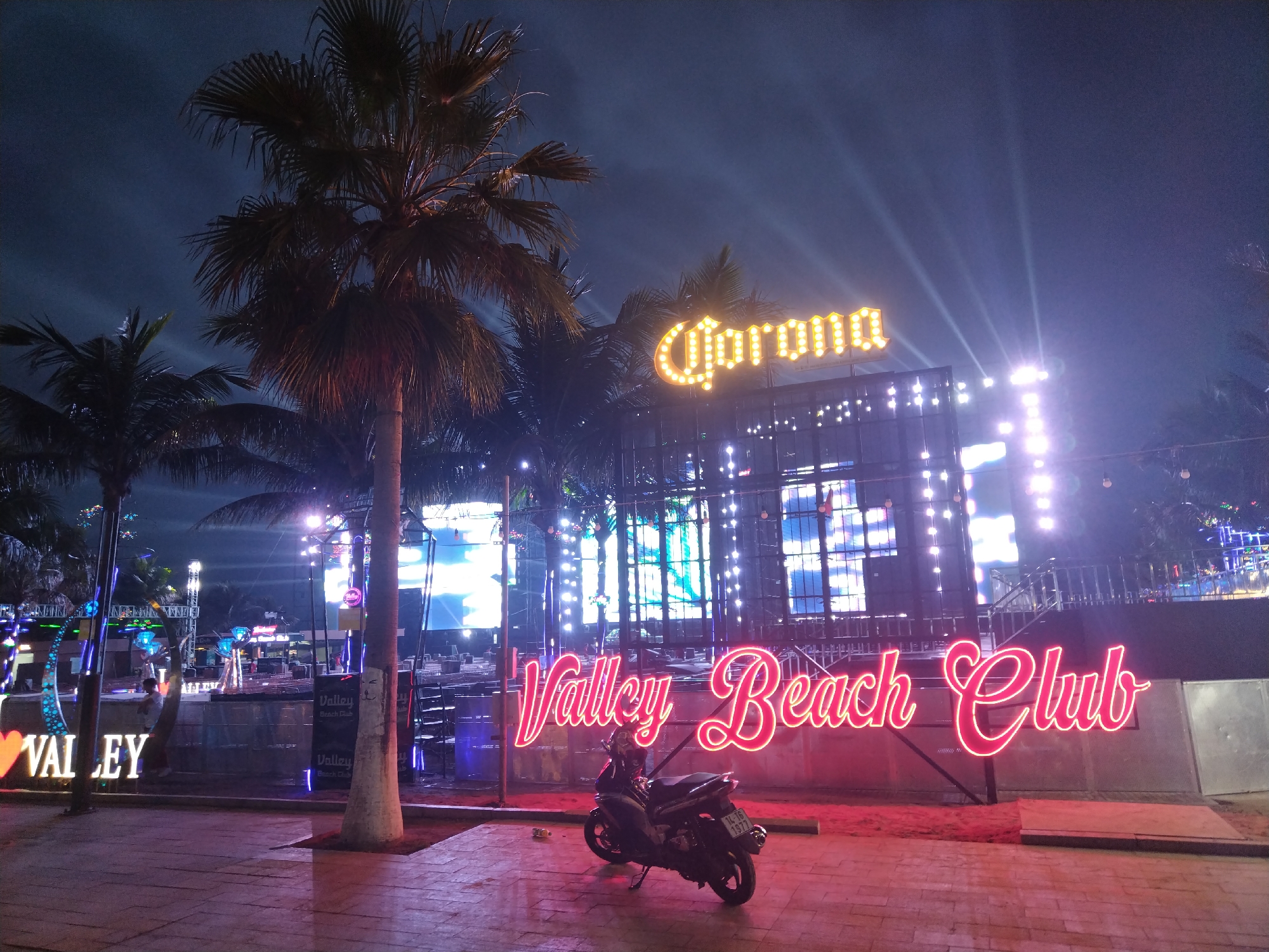 The image presents a vibrant nighttime scene of an outdoor entertainment venue, identified as the "Valley Beach Club," located in Bai Chay Ward, Vietnam. In the foreground, a large sign with pink and red neon-style lights spells out "Valley Beach Club." In front of this sign, a dark scooter or motorbike is parked on a paved area, with its Vietnamese license plate "14-L6 197.7" clearly visible.
Behind the club's main sign, a multi-level structure is brightly lit with numerous lights, some casting powerful beams into the dark night sky. Above this structure, a prominent yellow marquee-style sign displays the word "Corona." Several tall palm trees are scattered throughout the scene, their silhouettes visible against the glowing lights and the dark sky, reinforcing the tropical beach club setting.
To the far left, a partially visible white sign reads "I ❤️ Valley." A small, indistinct figure can be seen near this sign. Other smaller banners inside the club also feature the text "Valley Beach Club." The overall atmosphere is lively and festive, indicative of a place for evening entertainment, though no large crowds are present. The strong artificial lighting dominates the scene, highlighting the club's features against the dark backdrop.