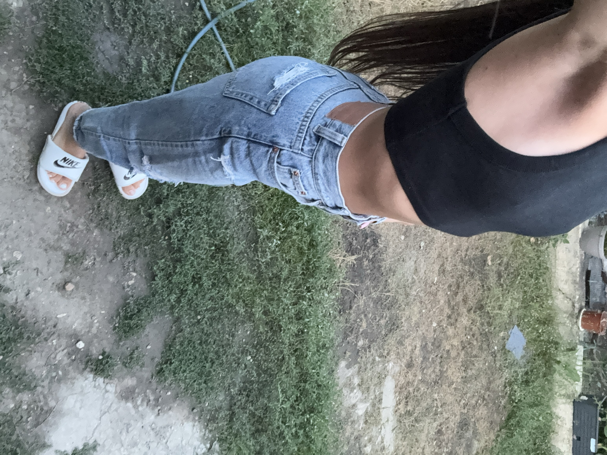 A person, viewed from the side, is standing outdoors on a patch of dry and green grass. They are wearing a black crop top and light-wash ripped jeans. White Nike slides are on their feet. In the background, there are potted plants and a rough concrete structure. The scene appears to be in a backyard or garden area. The provided location of Basarabeasca, Moldova, is not discernible from the visual information in the image.