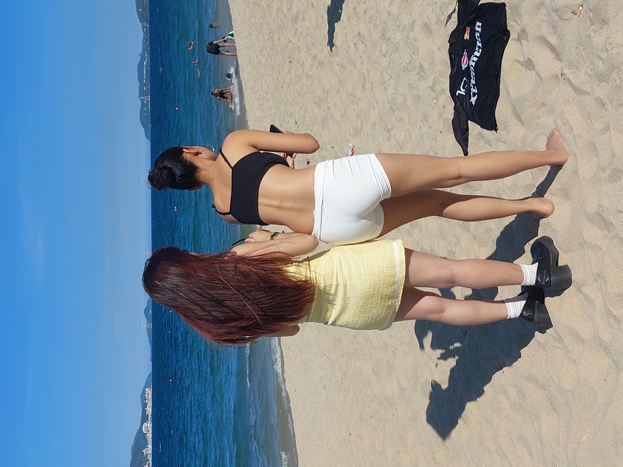 Two women stand on a sandy beach in Nha Trang Ward, Vietnam, with the blue ocean and a distant coastline under a clear sky. The woman on the left has long, dark red hair and wears a textured yellow mini-dress and black platform loafers with white socks. The woman on the right has her dark hair in a bun, wears a black sports bra and white athletic shorts, and holds a mobile phone. Several people are visible in the ocean in the background, some swimming and others in shallow water. A black t-shirt with white lettering lies on the sand near the woman on the right. The environment includes the sandy beach, gentle waves lapping the shore, and hills or mountains in the distance with some structures on them, possibly indicating the city of Nha Trang.