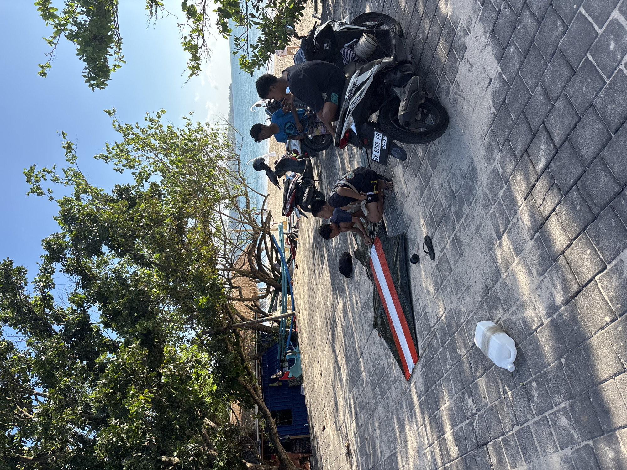 The image captures a bright, sunny day in Kuta, Indonesia, along a paved pathway bordering a sandy beach and the ocean. Several young men are gathered, primarily engaged in preparing what appears to be a large kite.
In the foreground, two individuals are crouched on the grey paver blocks, focused on a long, flat object laid out on the ground. This object has a prominent design featuring a wide red stripe over a white stripe, resembling the colors of the Indonesian flag, strongly suggesting it is a traditional kite. Nearby, a pair of black flip-flops and a white plastic jug are resting on the pavement.
Further back, two other young men stand next to parked motorbikes. One motorbike clearly displays an Indonesian license plate, "DK 6268 FR", with "06-29" possibly indicating a registration date. The group appears focused on their activities, with an air of concentration.
The setting is characterized by lush green trees providing shade along the pathway, their leaves brightly lit by the sun. Beyond the paved area, a sandy beach transitions into the calm, blue waters of the ocean, stretching under a clear, bright sky. Strong shadows cast by the trees and motorbikes indicate it is likely midday. Hints of distant buildings can be seen through the foliage. The scene depicts a common pastime in coastal areas, with the individuals engrossed in readying their kite for flight near the sea.