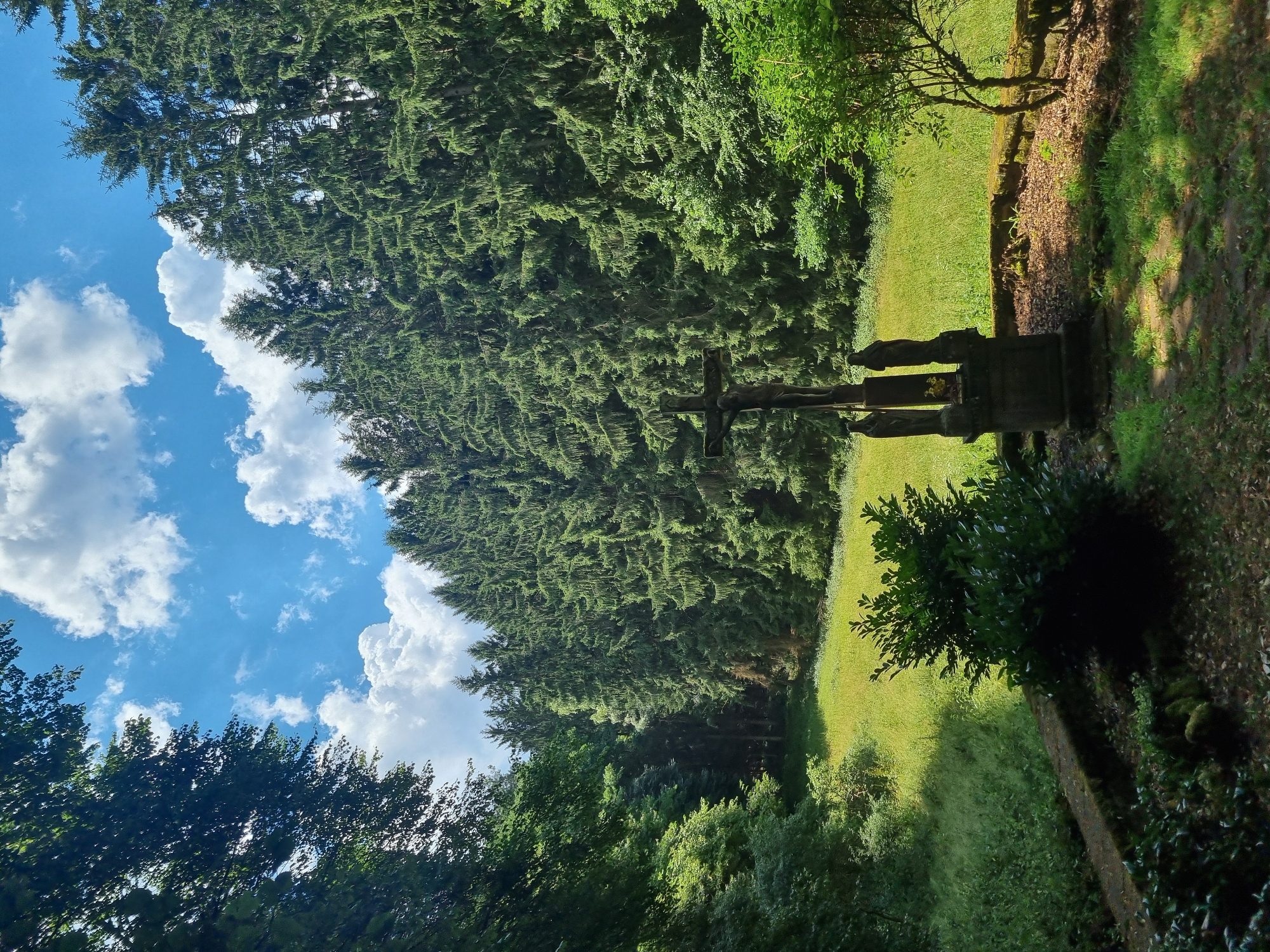 This media file depicts a scenic outdoor setting in Dahn, Germany. The primary subjects are a stone crucifix with figures, situated amidst a lush green landscape. Towering pine trees form a dense forest bordering a sunlit grassy area. The sky above is a vibrant blue, dotted with fluffy white clouds, suggesting fair weather. The lighting indicates it is daytime, likely late morning or early afternoon, with sunlight casting distinct shadows. The overall mood is serene and contemplative, emphasizing the natural beauty of the location and the presence of the religious monument. There is no visible text in the image.