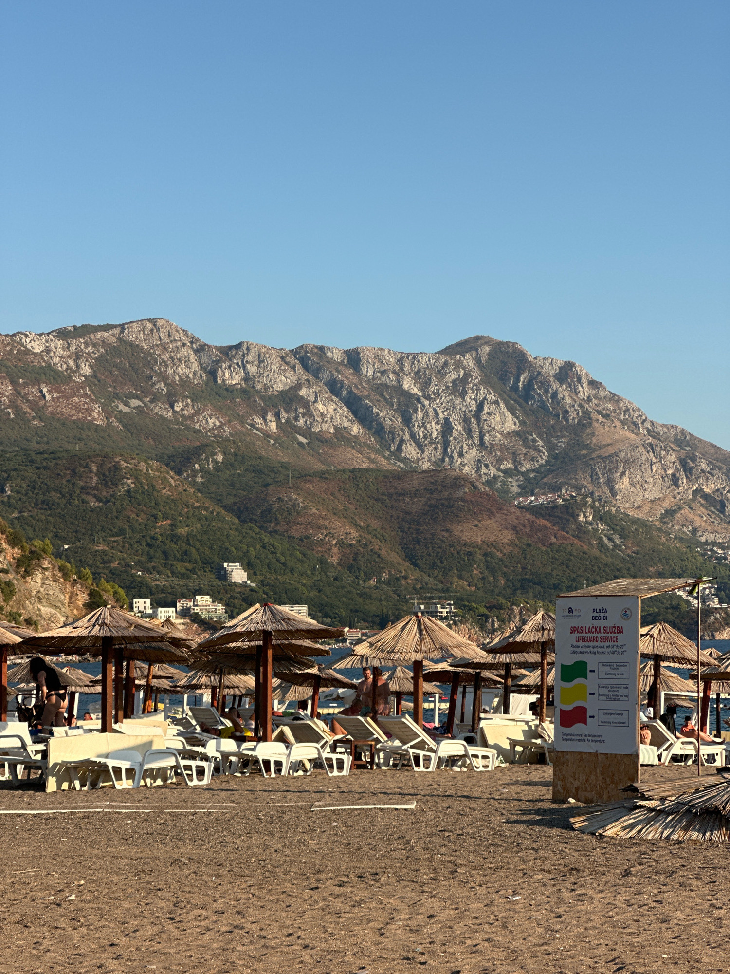In Boreti, Montenegro, a sandy beach is dotted with numerous thatched-roof umbrellas and white lounge chairs, many unoccupied. A signpost displays the "Lifeguard Service" information with colored flags indicating swimming conditions. In the background, lush green hills transition into rugged, rocky mountains under a clear blue sky. A few people are visible on the beach, some sitting in chairs and others walking. Buildings are nestled into the hillsides. The scene captures a tranquil day at the beach.