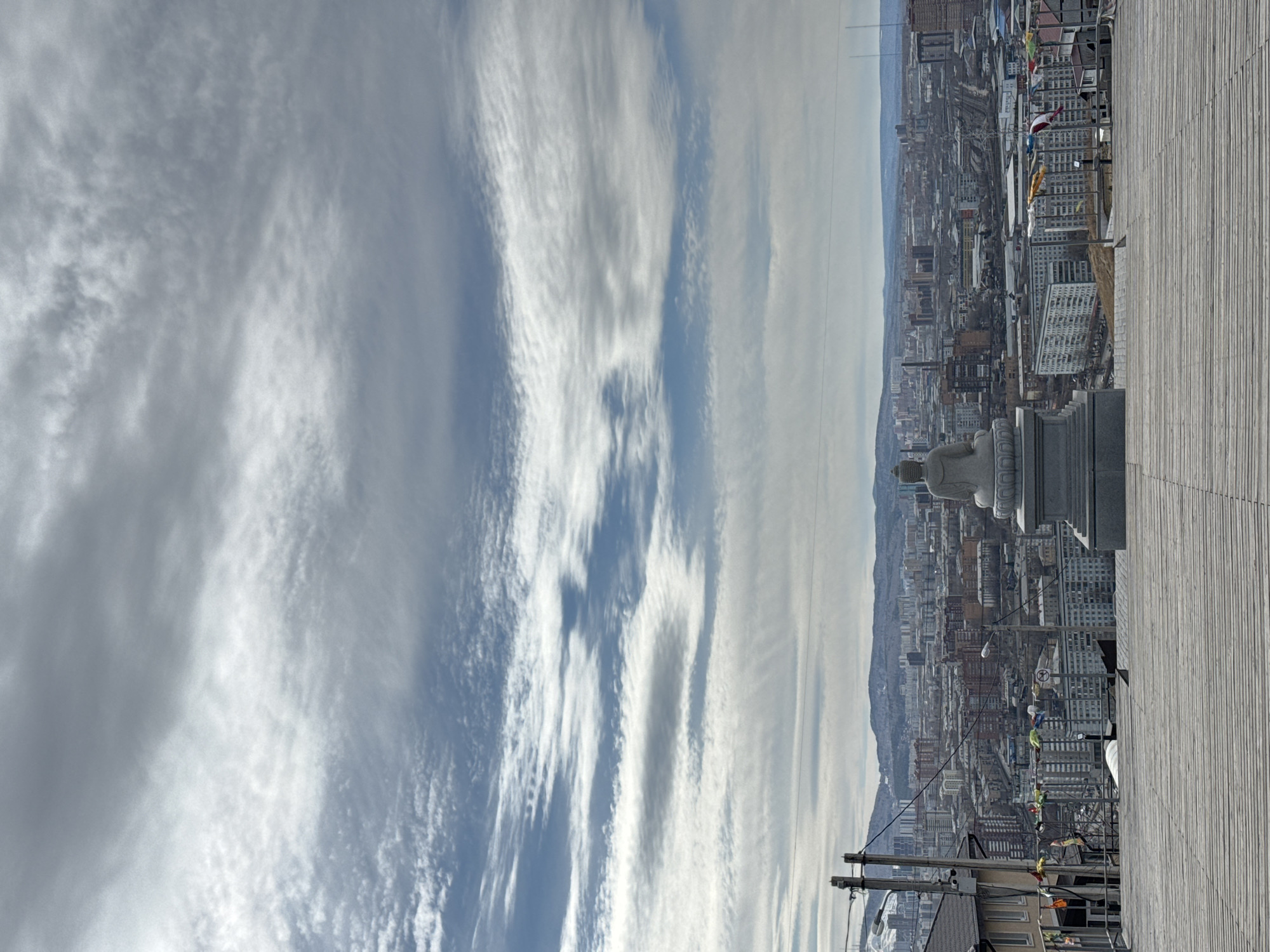 The image depicts a high-angle view of Krasnoyarsk, Russia, likely taken from the Paraskeva Pyatnitsa Chapel observation point. The foreground includes a section of a wooden deck or boardwalk, with a portion of a stone monument visible at the edge. The city stretches into the distance, with buildings of varying heights densely packed together. The backdrop consists of a mountain range partially obscured by a hazy sky. The sky itself is filled with streaky, textured clouds. A utility pole with wires is visible in the lower part of the frame. The overall impression is one of a panoramic city view under a cloudy sky.