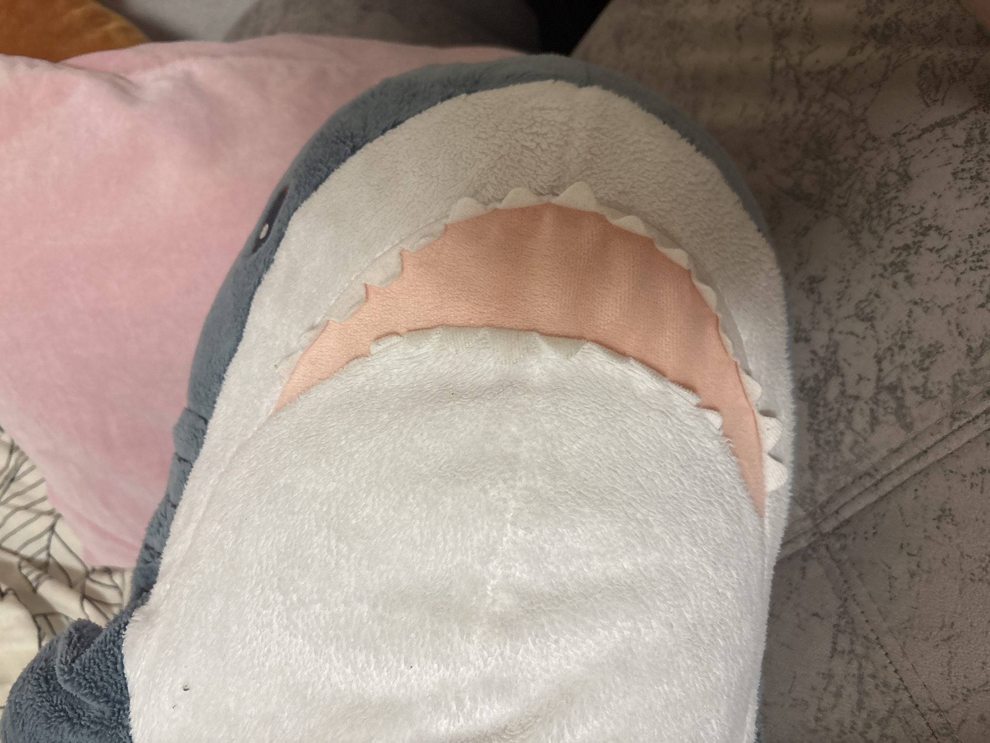 The image shows a close-up of a plush shark toy, with its white belly and pink mouth with white teeth clearly visible. The shark is resting on a textured gray surface, likely a sofa or chair. In the background, out of focus, are parts of other soft furnishings, including a pink pillow and a striped blanket. There are no people or any other distinguishing features visible that would indicate a specific location in Kazan, Russia, or any other city.