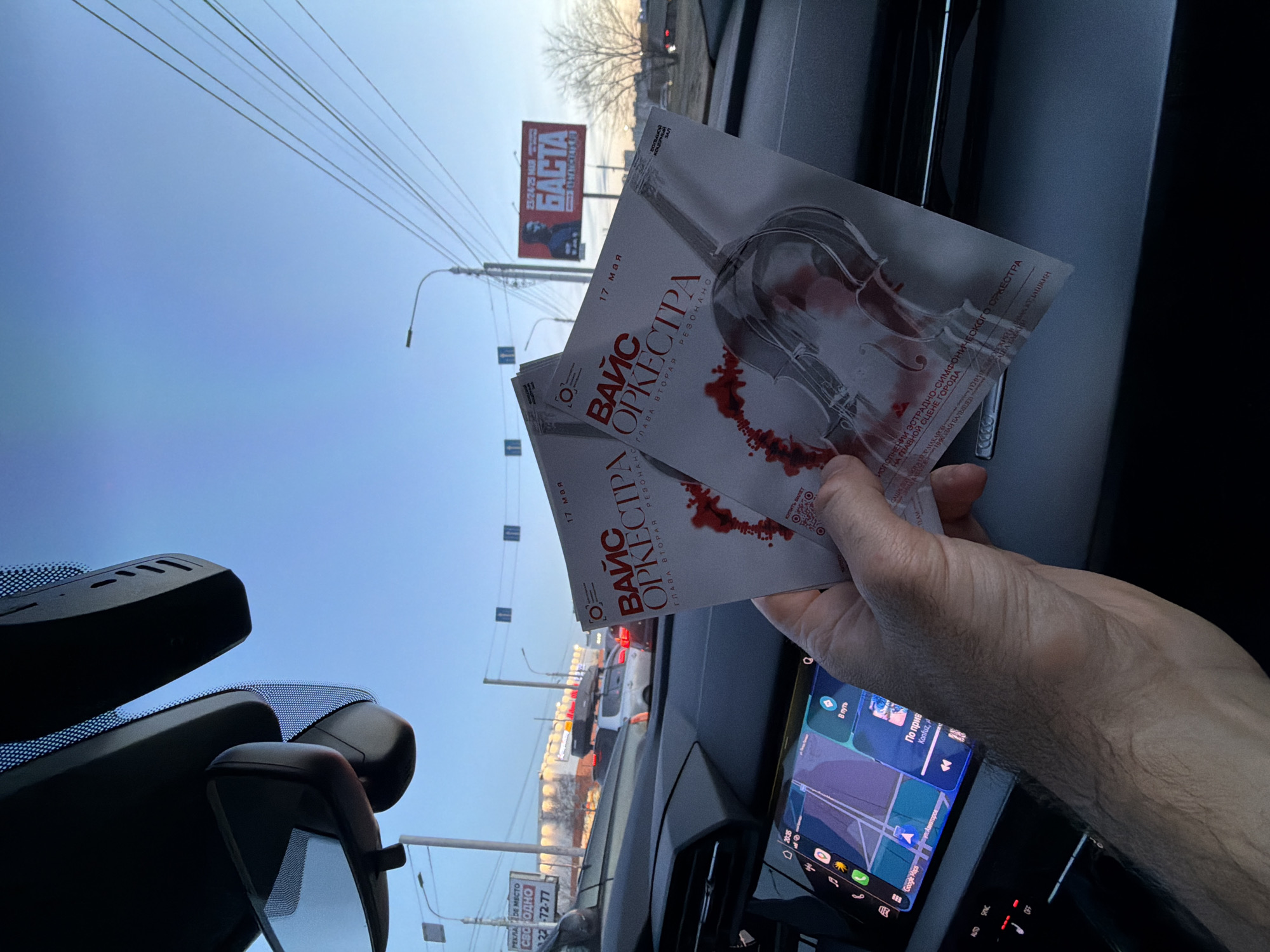 The image captures a first-person perspective from inside a car, looking out at a street scene in Krasnoyarsk, Russia. In the foreground, a person's right hand holds two flyers or tickets for an event. The tickets are for "ВАЙС ОРКЕСТРА" (VICE ORCHESTRA), "ГЛАВА ВТОРАЯ РЕЗОНАНС" (CHAPTER TWO RESONANCE), scheduled for "17 мая" (May 17), and feature an image of a violin with red, blood-like splatter. Text indicates it's a "POP SYMPHONIC ORCHESTRA" performance "ON THE MAIN STAGE OF THE CITY."
Through the car's windshield, a street with light traffic is visible under a clear, bright blue sky, suggesting late afternoon or early evening. Power lines crisscross the upper part of the sky. In the middle distance, a large billboard prominently displays "БАСТА" (Basta, a Russian rapper) and dates "23/24/25 МАЯ" (May 23/24/25) for an event at the "GRAND CONCERT HALL." A smaller sign lower on the left advertises an "ADVERTISING PLACE AVAILABLE" with a phone number.
Inside the car, parts of the dashboard, rearview mirror, and an infotainment screen are visible. The screen shows "Google Maps" with Russian text "В путь" (On the way) and "По прив," likely indicating a song playing or recently played ("По привкусу" by Konfuz). Car controls with labels like "AUTO," "SINC," and "OFF" are also present. The scene implies the person is in transit, possibly heading to or from an event, given the tickets and the navigation on the car's screen.