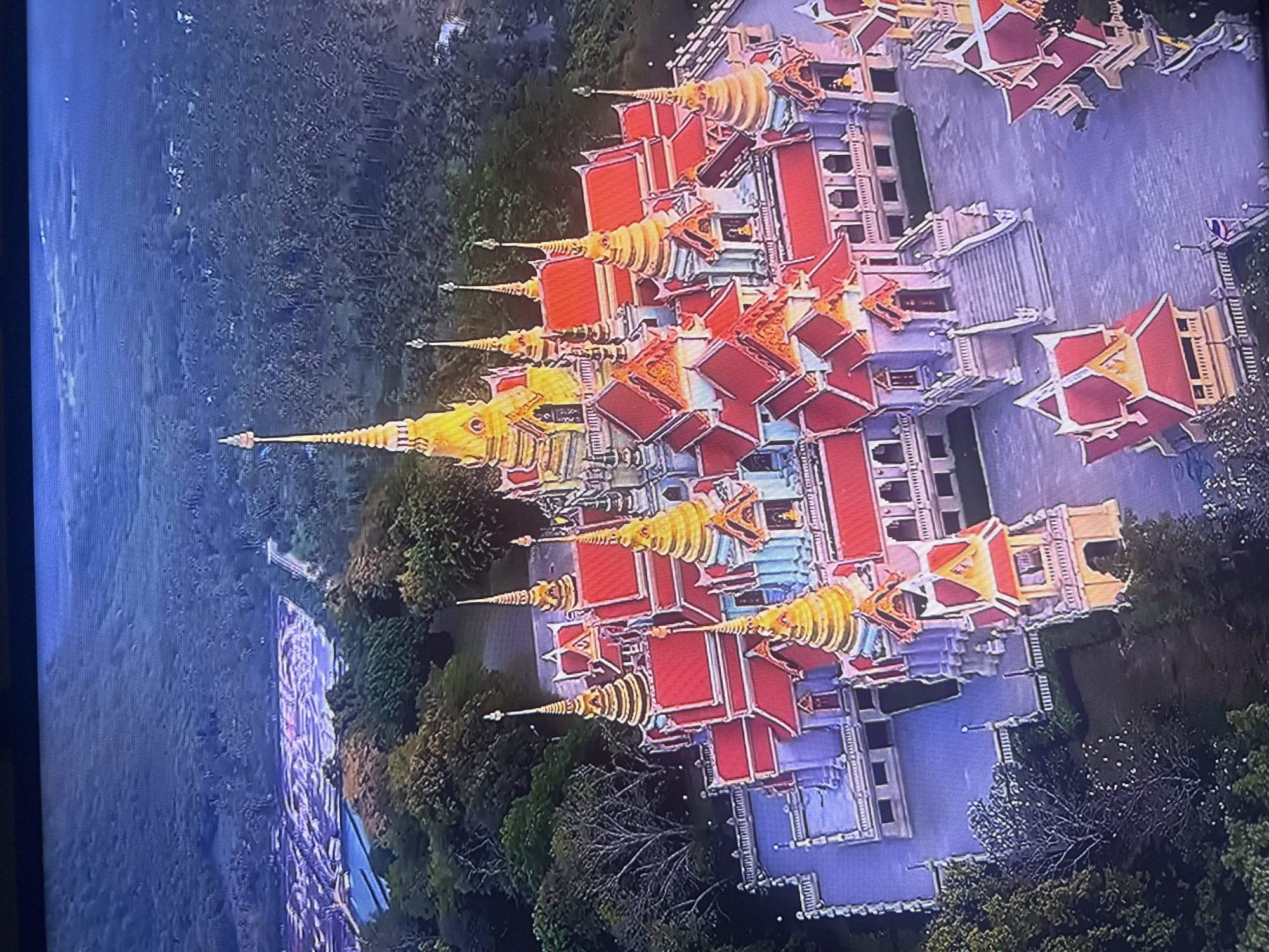 The image shows an aerial view of a building complex, likely a temple or palace, with intricate architecture featuring red roofs and golden spire-like structures. It is surrounded by lush greenery, possibly trees or a forest. A portion of what may be a city or populated area is visible on the left side of the image. The complex features detailed architectural elements and appears well-maintained. There is no location of Varna, Bulgaria in the image.