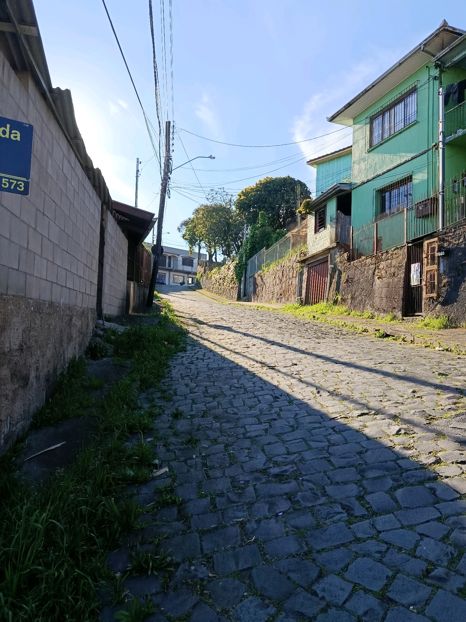 The image shows a cobblestone street in Caxias do Sul, Brazil, ascending a gentle slope. On the left, a weathered, light-colored brick wall borders the street, with a blue street sign that reads "da" at the top and "573" below. Tall grass grows along the base of the wall. On the right side of the street, two multi-story houses with green painted facades are visible. The houses have barred windows and a metal fence in front of them. A wooden gate is open on the ground floor of the house closer to the foreground, revealing a dark entryway. Overhead, telephone wires stretch across the bright blue sky, casting long shadows across the uneven cobblestones, indicating a sunny day. The street appears quiet and residential, with no people or vehicles present in the frame. The overall impression is one of an ordinary urban street scene bathed in the warm light of the sun.