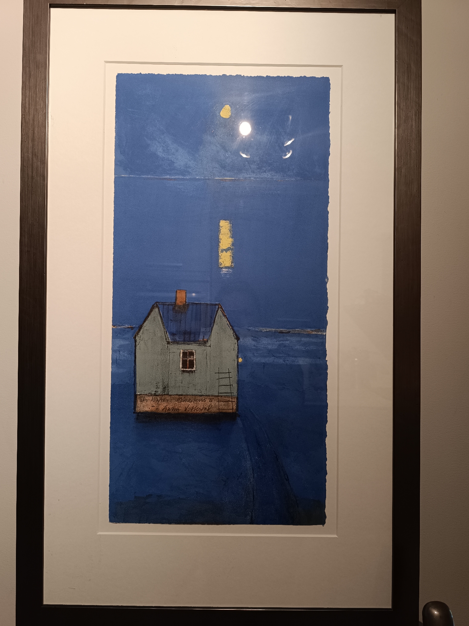 This image displays a framed artwork featuring a solitary, pale blue house with a reddish-brown chimney and a small window, situated on a platform that floats on dark blue water. The artwork is matted with a thick white border and framed in dark wood. In the background, a faint yellow rectangular shape appears on the water, and a circular, glowing yellow orb is visible in the deep blue sky, possibly representing the moon or a light source. The artwork is signed in cursive script at the bottom, but the writing is indistinct. The scene is rendered in a stylized, somewhat abstract manner. The photograph of the artwork is taken from a frontal perspective, with a slight downward angle. The background wall is a neutral, light beige color. The artwork is likely a print or painting, and the lighting in the room creates a subtle reflection on the glass covering the artwork.
The artwork depicts a scene that could be inspired by the coastal environment of Hovden, Norway, a location known for its maritime character and the presence of water bodies. The solitary house and the tranquil water evoke a sense of isolation and peacefulness that might be associated with a remote coastal dwelling in Norway.