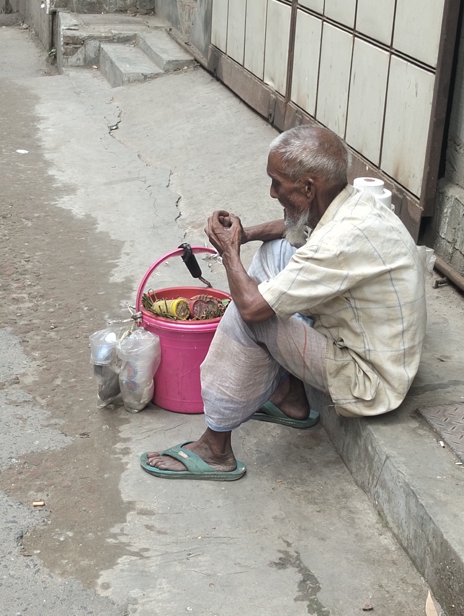 An elderly man sits cross-legged on a concrete step next to a metal gate in Dhaka, Bangladesh. He has grey hair and a white beard and is wearing a beige plaid shirt, light blue pants, and green flip-flops. A pink plastic bucket sits beside him, filled with items, including what appears to be betel nut and leaves, with plastic bags containing cups. A cigarette butt lies on the ground nearby. The sidewalk is cracked and stained.