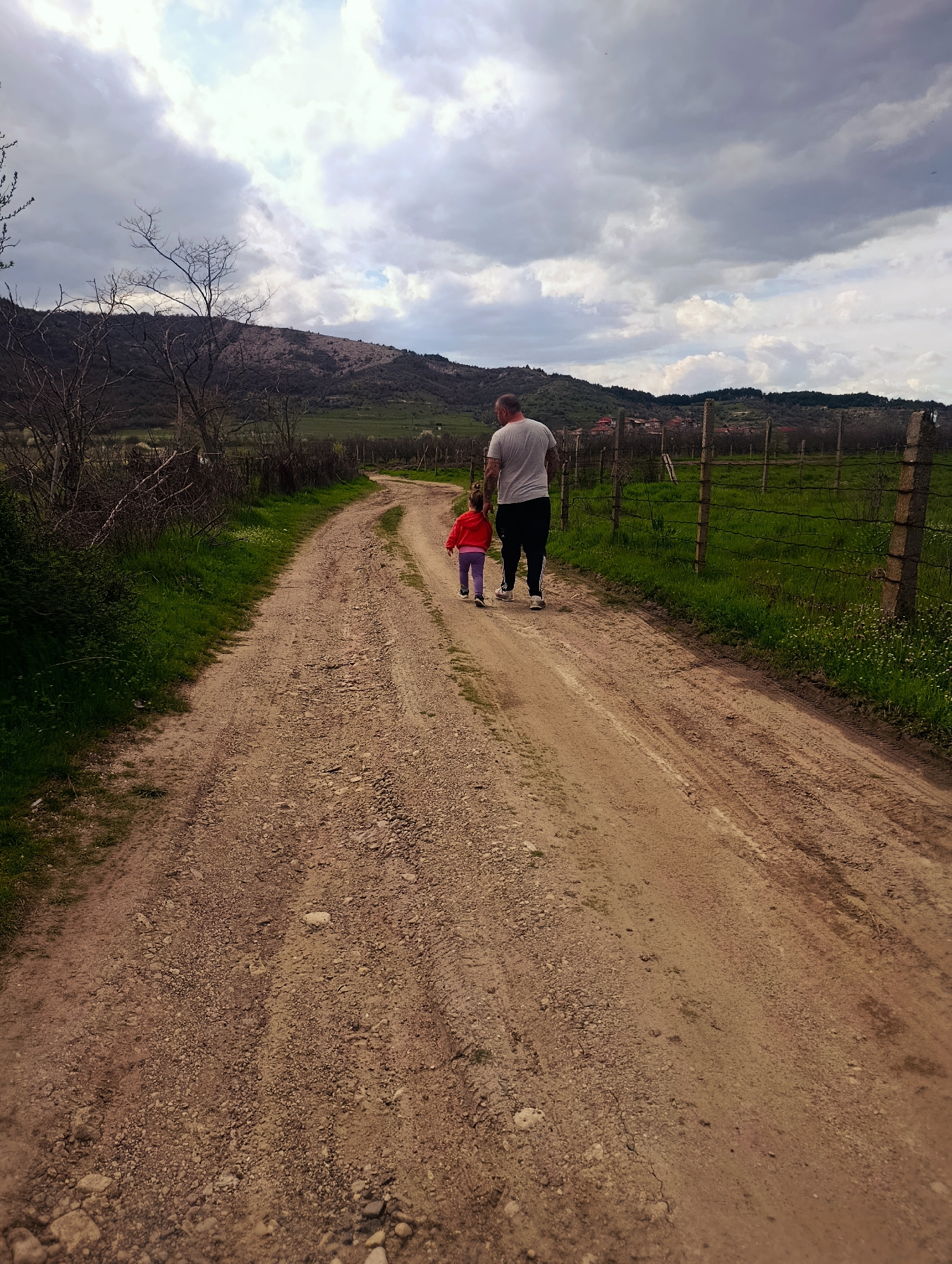 A medium shot depicts a father and child walking away from the camera on a dirt road. The location is Shiroko pole, Bulgaria. The father is wearing a light grey t-shirt and black pants, and appears to have tattoos on his back. The child is wearing a red jacket and purple pants. They are walking away from the viewer. The dirt road is rocky and uneven. On either side of the road is green grass and vegetation. A barbed wire fence runs along the right side of the road. In the background, there are rolling hills and a cloudy sky. The overall atmosphere is rural and overcast.