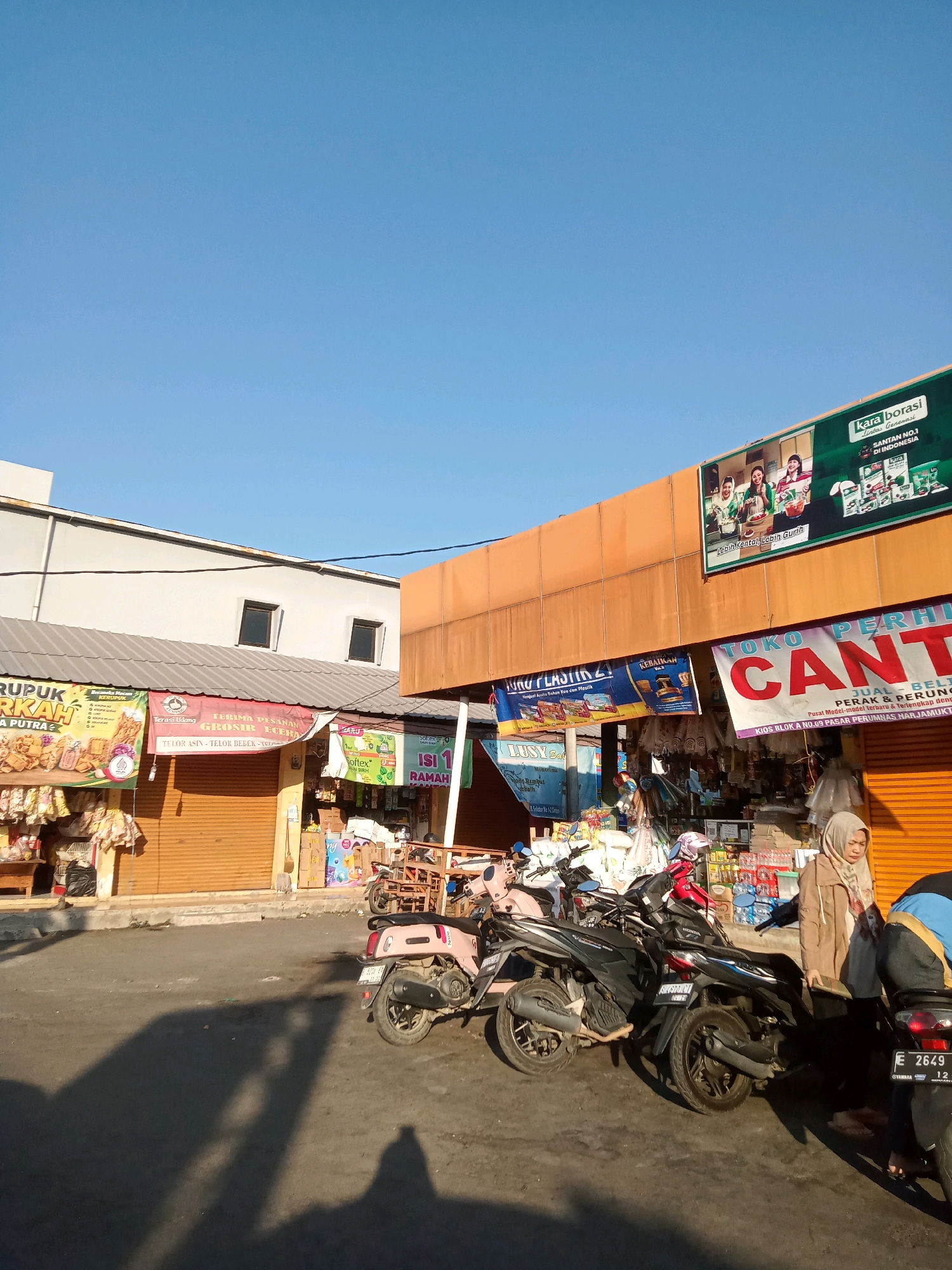 This is a daytime, eye-level, outdoor, street-level photograph taken in Cirebon, Indonesia. The image depicts a row of small shops, likely in a market or commercial area, with their metal shutters partially or fully closed. The architecture suggests a modest, utilitarian commercial zone.
In the foreground, several motorcycles are parked on an asphalt surface, casting long shadows due to the low angle of the sun. Two people are visible: a woman wearing a light brown hijab and a beige jacket stands near a motorcycle, and another person, partially visible from behind and wearing a blue and yellow jacket, is also near a motorcycle.
The shops display various banners and signage, some in Indonesian. One large banner on an orange facade reads "TOKO PERHI CANT," with additional text below that is partially obscured. Another banner above a shop entrance features images of products and text including "KARAI BORASI" and "SANTAN NO.1 DI INDONESIA." Other signs indicate products like "KERUPUK" and "PLASTIK." Merchandise is visible on display in front of some of the open shop fronts.
The sky is a clear, bright blue, indicating good weather. The overall scene conveys a sense of a typical day in a bustling, local commercial district.