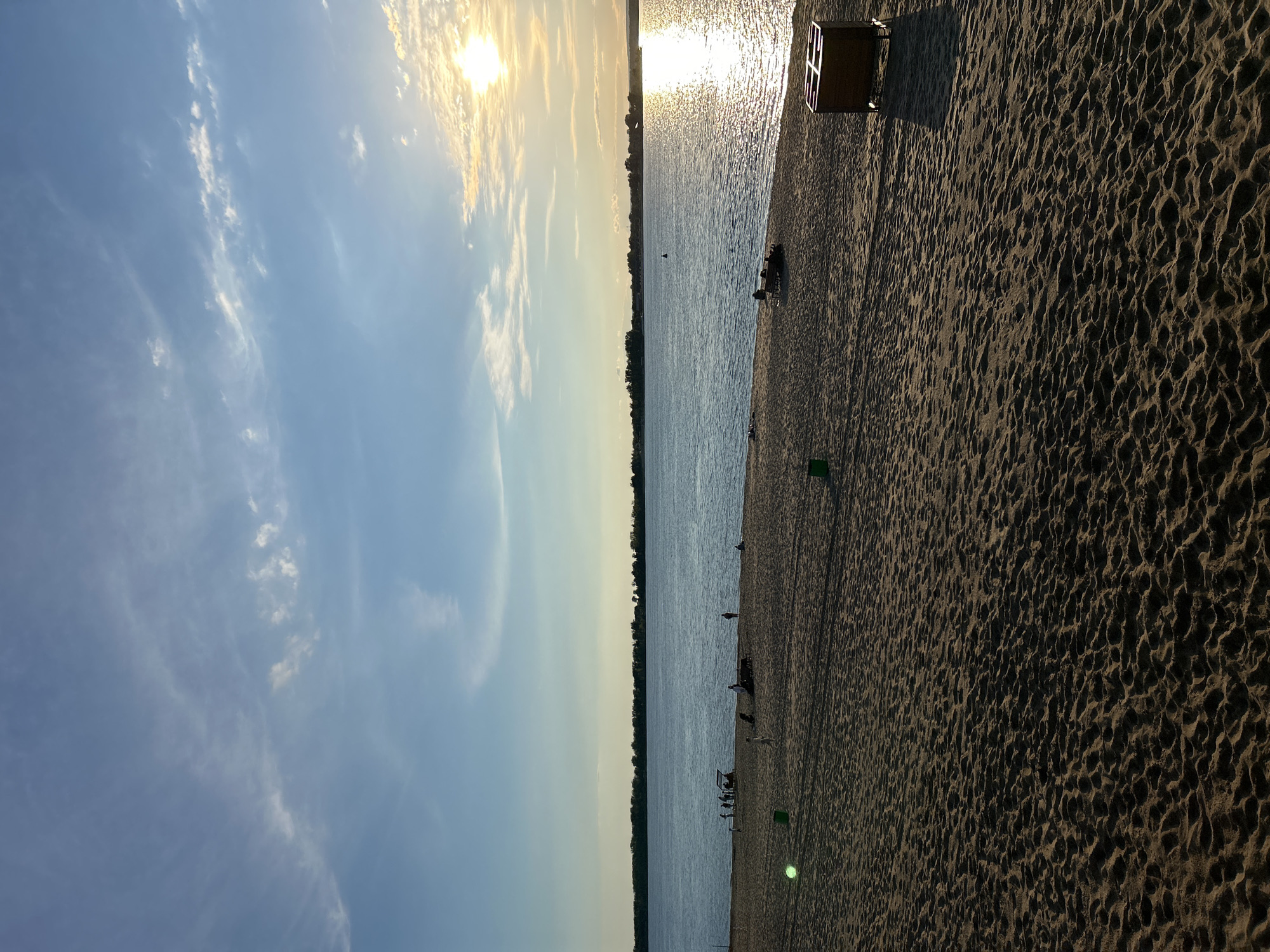 This is a photograph of a beach scene, oriented vertically. The sky occupies the left side of the image, displaying a bright, hazy sun low in the sky, casting a warm glow. Wispy white clouds are scattered across the pale blue sky. The right side of the image is dominated by a sandy beach, with distinct patterns of footprints and ripples in the sand.
The water of a large body of water, likely a river or lake, is visible in the center, reflecting the sunlight and appearing textured with small waves. Along the shoreline, several people are present. Some are standing or walking, while others appear to be sitting or relaxing. There are also a few small objects scattered on the sand, including what appears to be a wooden structure or piece of furniture near the top of the sand area and green trash receptacles. The overall impression is of a late afternoon or early evening at a public beach. The lighting suggests a warm, possibly humid, day. The presence of trees along the distant horizon indicates a natural or park-like setting. The image evokes a sense of leisure and calm. No visible text is present in the photograph.