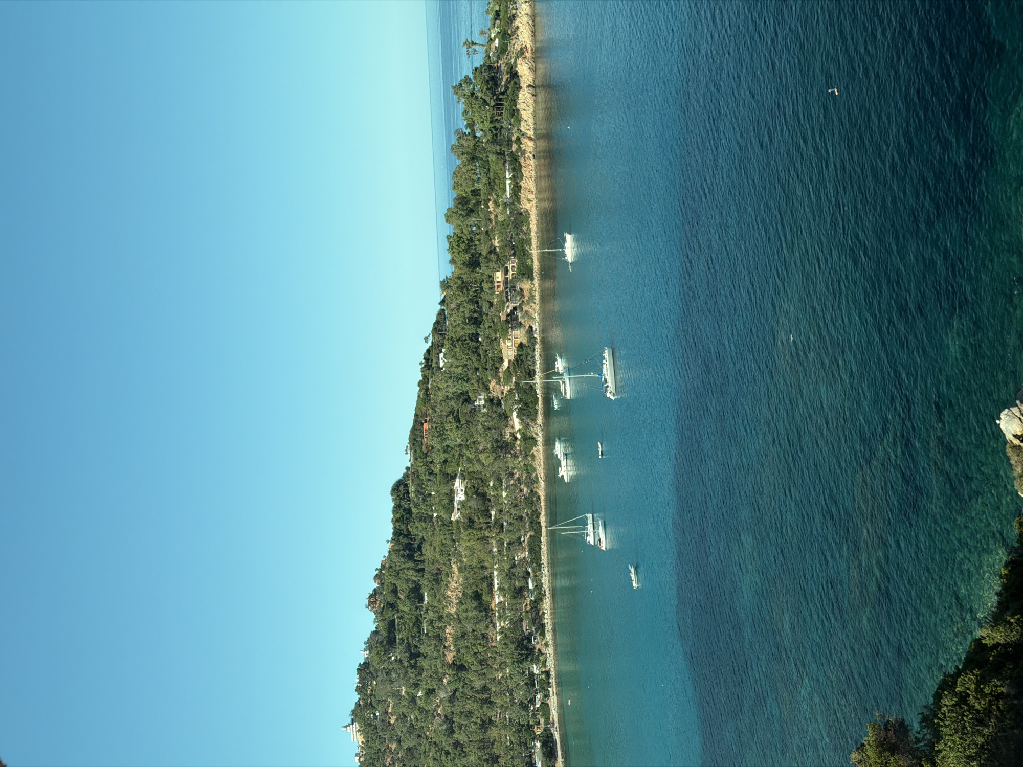 This is a high-angle, long shot of the coastline of Tortolì, Italy. The foreground is dominated by deep blue-green water with visible seabed in shallower areas. Several sailboats are anchored in the bay, with one larger white sailboat prominently featured. The middle ground shows a tree-covered hillside dotted with buildings, some appearing to be villas. The background consists of the clear blue sky meeting the calm sea. A single person is visible in the water in the lower right quadrant of the image.