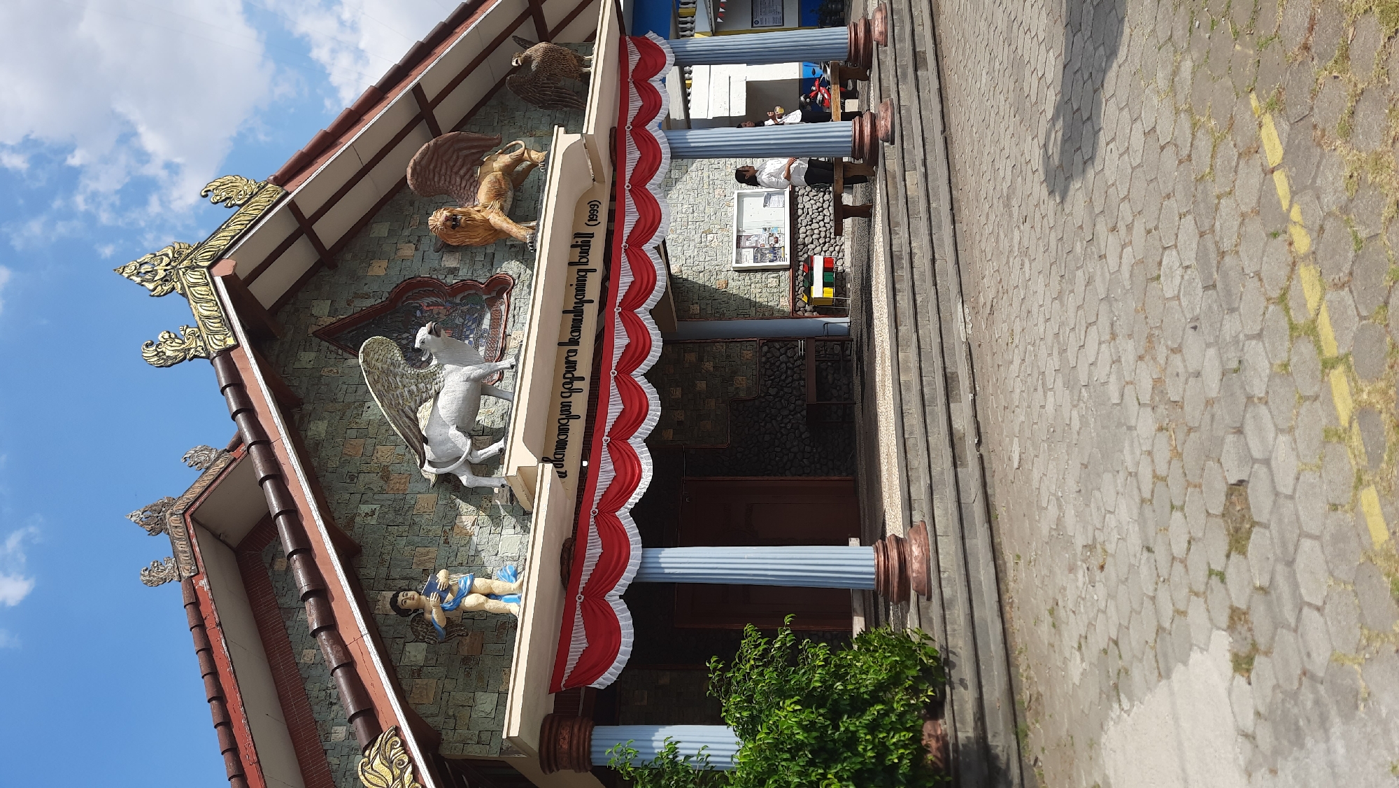 The image shows the exterior of a building with an inscription "slamun gapura kamulyaning budil (1999)". The building has a textured stone facade and a red tiled roof adorned with golden, ornate decorations. Statues of a winged angel, a winged bull, a winged lion, and a bird are placed on the facade. A red and white valance decorates the entrance area, which is supported by blue and brown columns. Stone steps lead up to the entrance, and a lush green bush is visible to the left. Paved hexagonal tiles form the ground in front of the building, with a yellow line marking a path. Two women are standing near the entrance, one facing left and the other looking towards the right. One woman is wearing a white shirt and dark pants, and the other is wearing a dark top and pants. Sunlight casts shadows on the scene, and a blue sky with scattered clouds is visible above. The building is situated in Nanggulan, Indonesia.