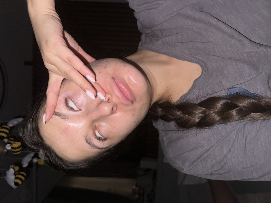 The image depicts a close-up, eye-level view of a single individual, likely a female, indoors in the city of Unknown, Unknown. The subject, appearing to be a young adult, has dark brown hair styled in a single braid resting over her left shoulder and chest. She is wearing a grey v-neck t-shirt. Her face is oriented slightly to her right, with her eyes rolled upwards, revealing only the white sclera. Her lips are parted, appearing full and glossy, with a slight pouting expression. Her skin exhibits some visible blemishes and appears shiny, possibly from moisture or a product. The individual's left hand is raised, with fingers spread, contacting her face; the ring finger touches the bridge of her nose, and the middle finger is near her left eye. Her fingernails are manicured with light-colored polish and small glitter accents. In the upper left background, a decoration consisting of three plush or yarn-made bees (yellow and black stripes with white wings) is visible hanging. The background is dark, out of focus, and suggests a dimly lit indoor environment.