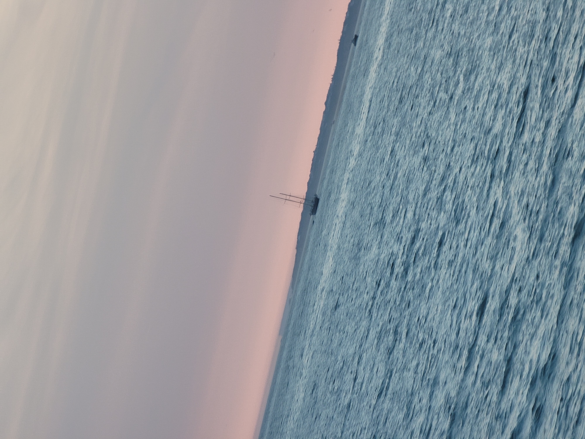 The image shows a body of water with a faint pinkish-purple sky. Two sailboats are visible in the distance on the water. One sailboat, a larger schooner, is on the left side of the image. A smaller boat is on the right side. The water has gentle ripples. The scene appears to be a coastal view.