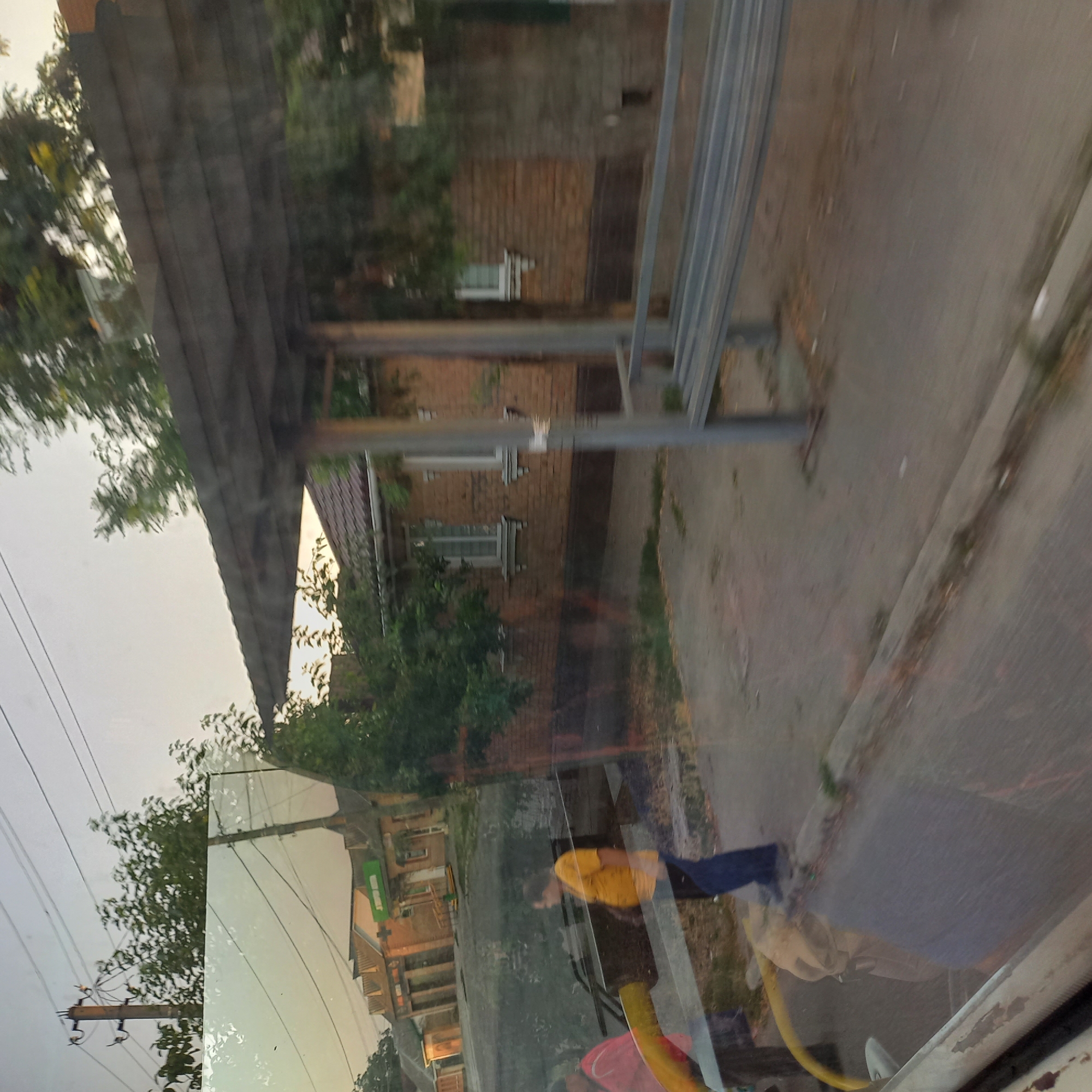 The image shows a street scene in Kropyvnytskyi, Ukraine, likely captured through a window. In the foreground, a person wearing a yellow t-shirt and dark pants is visible, appearing to walk. Another person in a red jacket is partially visible to the left. Behind them, the reflection on the window shows a building with a green sign that reads "SUPER MARKET". Further back, a bus stop shelter with a wooden roof and metal supports is present, with a metal bench underneath. Beyond the shelter, a brick building with multiple windows and a tiled roof is visible. Trees and electrical poles line the street. The overall lighting suggests it is either early morning or late afternoon. The scene appears to be an ordinary street with commercial establishments and public amenities.