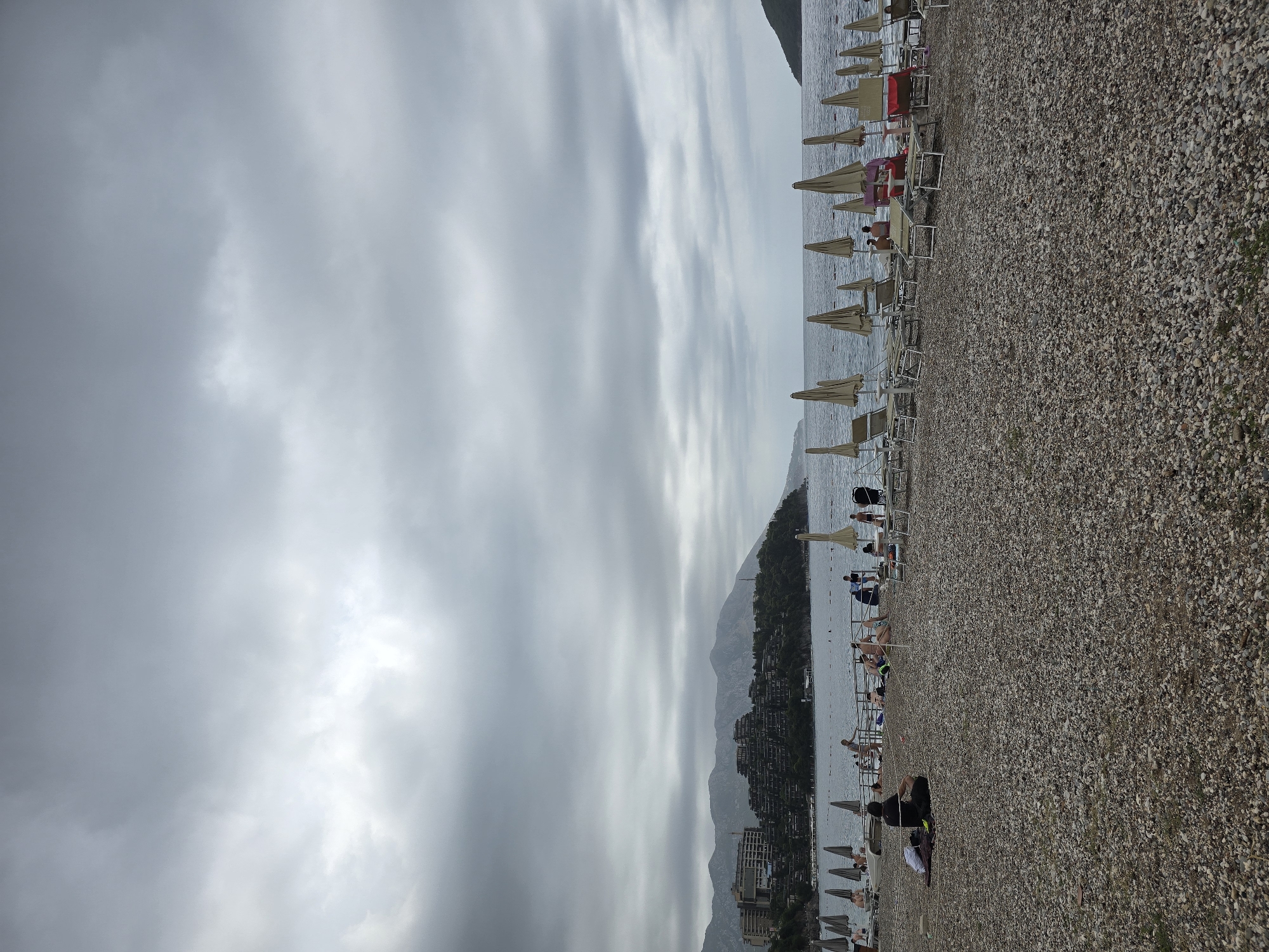 The image captures a cloudy day at a pebble beach in Budva, Montenegro. In the foreground, the beach is covered with small, varied-colored pebbles. Several lounge chairs and closed beach umbrellas are set up. In the mid-ground, the sea is visible, with a gentle wave action. A line of more lounge chairs and umbrellas extends along the coastline. Several people are scattered across the beach and near the water, some sitting, others walking or in the water. In the background, a peninsula juts out into the sea, covered with dense green foliage and dotted with several multi-story buildings, likely hotels or residential complexes. These structures are built into the hillside, creating a tiered effect. The sky is overcast with thick, grey clouds, obscuring any direct sunlight and casting a muted light over the scene. The overall impression is a somewhat subdued atmosphere due to the cloudy weather.
