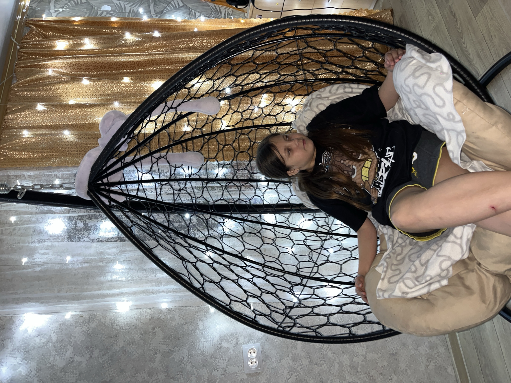 A young person lies in a black, net-like hanging chair. The chair is adorned with white lights. The walls are a light gray color. A brown and gold curtain is visible in the background. The person wears a black t-shirt and gray shorts. The individual is propped up by light-colored pillows and a patterned blanket. The scene takes place indoors in a room with an electrical outlet on the wall. The location is Novopokrovskaya, Russia.