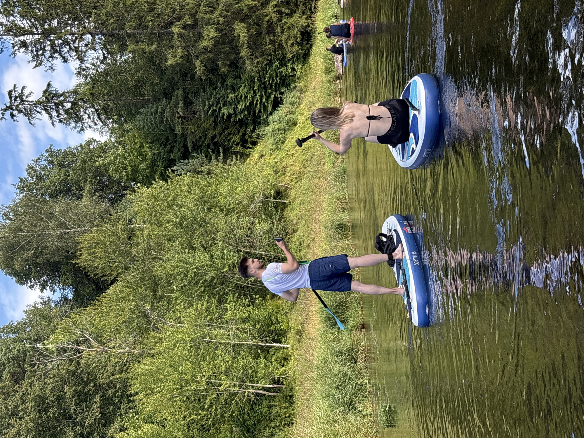 In the city of Starosiverskaya, Russia, a group of people are paddleboarding on a calm river. A man in a white t-shirt and blue shorts stands on a paddleboard, holding a paddle. A woman in a black bikini sits on another paddleboard, also holding a paddle. In the background, two more people are on paddleboards, and a dense forest lines the riverbank. The water reflects the surrounding trees and sky.