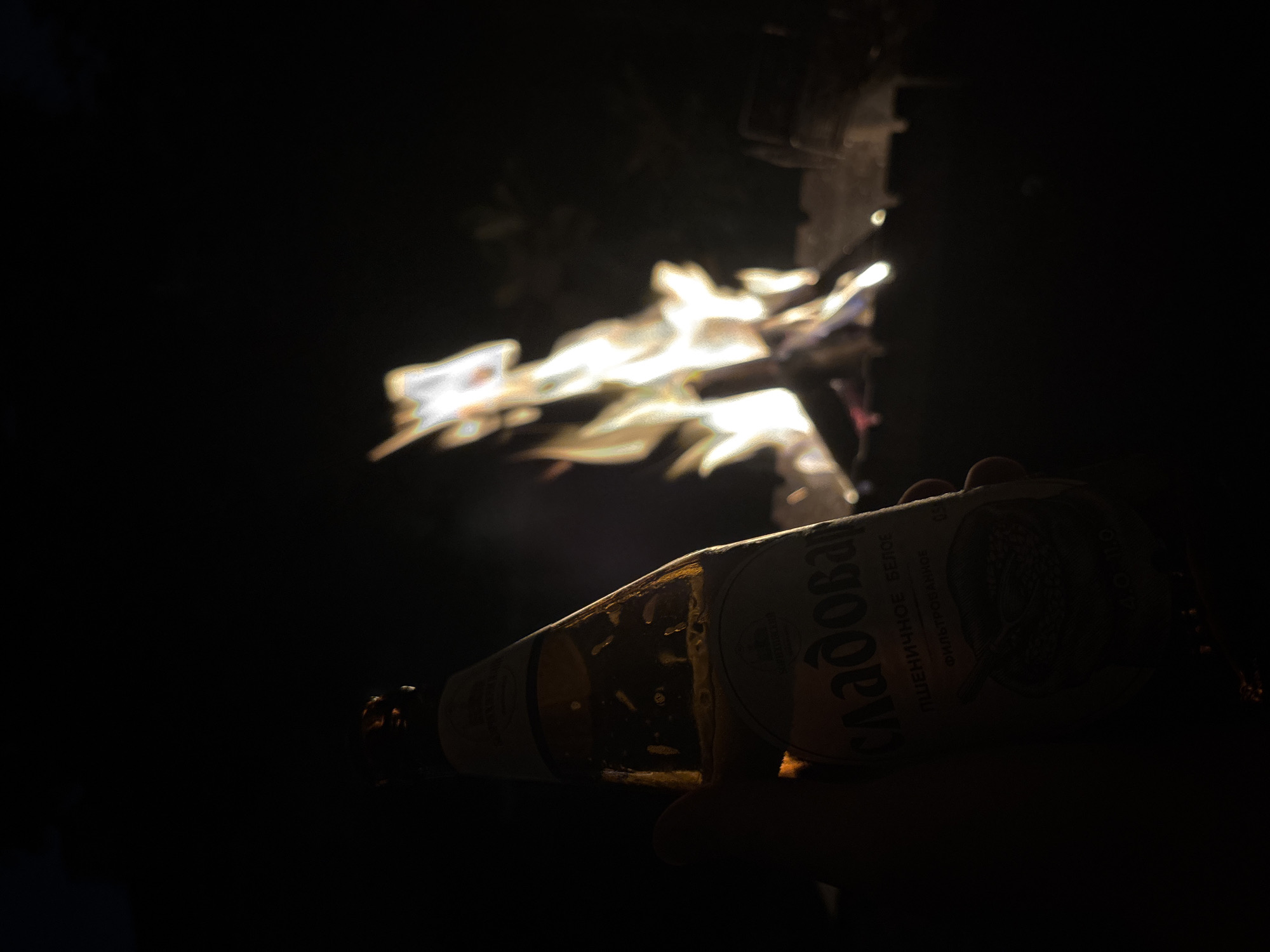 An outdoor scene at night features a hand holding a beer bottle in the foreground. The bottle, partially illuminated by ambient light, has visible condensation and a label with Cyrillic text, including the word "СЛАДОВАР". In the blurred background, a campfire burns with bright flames. The setting appears to be a park or backyard in Krasnodar, Russia, where a person is enjoying a drink by a fire.