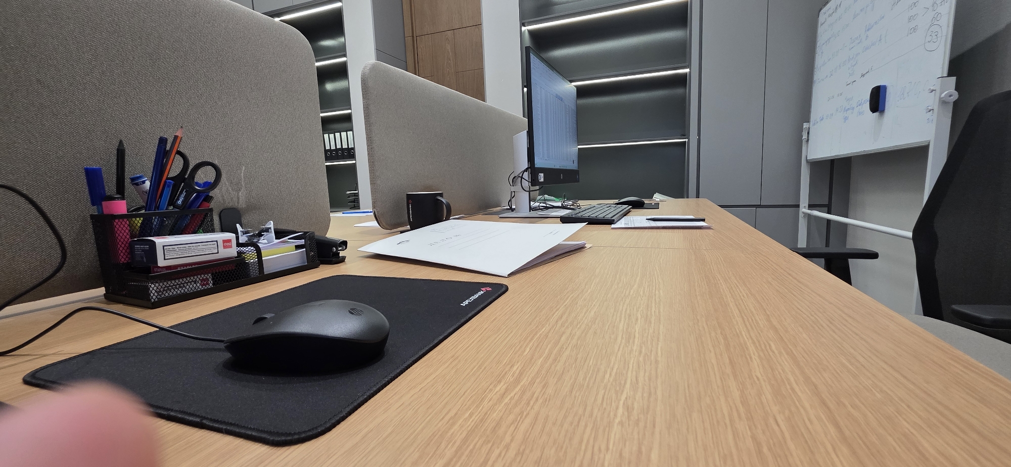 The image depicts a close-up, low-angle view of a wooden office desk. In the foreground, a black mousepad with a subtle logo is visible, with a black wireless mouse positioned on it. To the left of the mousepad, a portion of a person's finger is slightly blurred, suggesting the photo was taken by someone holding the camera.
The desk is cluttered with office supplies. To the left, a black wire mesh pen holder contains various writing instruments, including pencils and pens, and a pair of scissors. Below the pen holder, a black stapler and a small box with red and white labeling are stacked. Further back on the desk, a white document with text is open, and a black mug with a logo is placed behind it.
The background reveals part of an office cubicle with a grey fabric partition. A computer monitor displaying data is visible on a stand. Behind the monitor, shelving units with illuminated lights are present. To the right, a whiteboard on a stand contains handwritten notes and diagrams. A dark office chair is partially visible to the far right. The overall environment suggests a typical workspace in a modern office. The location of Tashkent, Uzbekistan, is not explicitly indicated by any visual cues in the image.