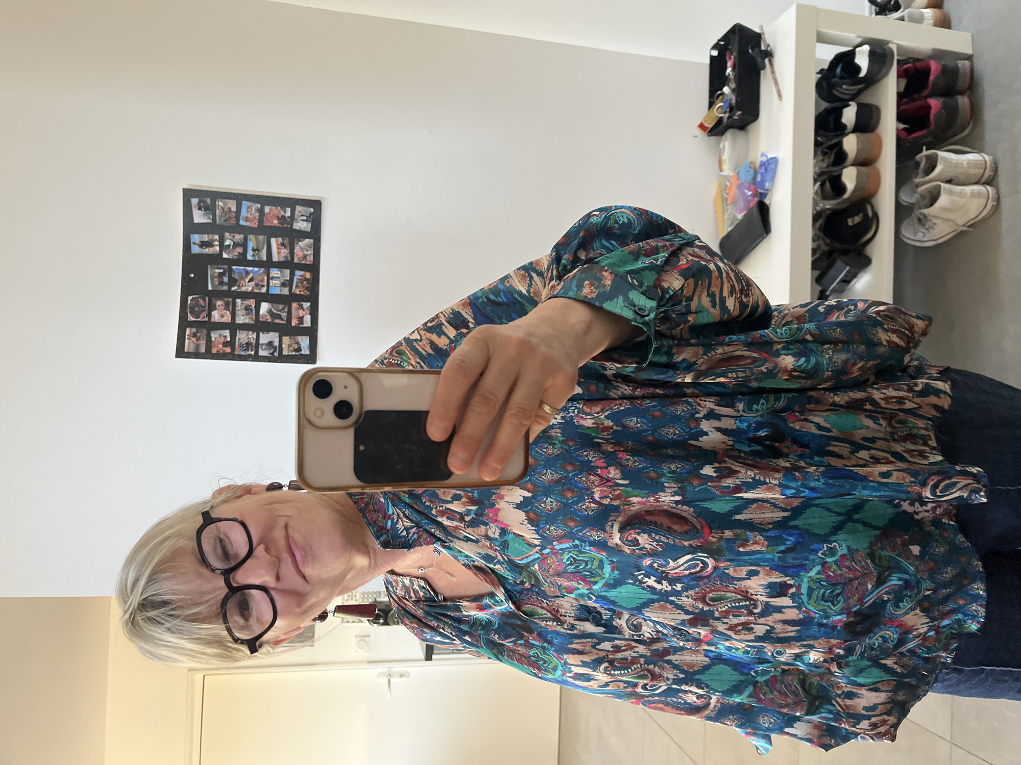 The image shows a woman taking a selfie in what appears to be her home. She is wearing glasses and a patterned blue and brown shirt. In the background, there is a white wall with a collage of small photos. To the right, a white shelving unit displays various pairs of shoes and a black container holding keys and other items. The exact location in Chambœuf, France, cannot be definitively determined from the image.