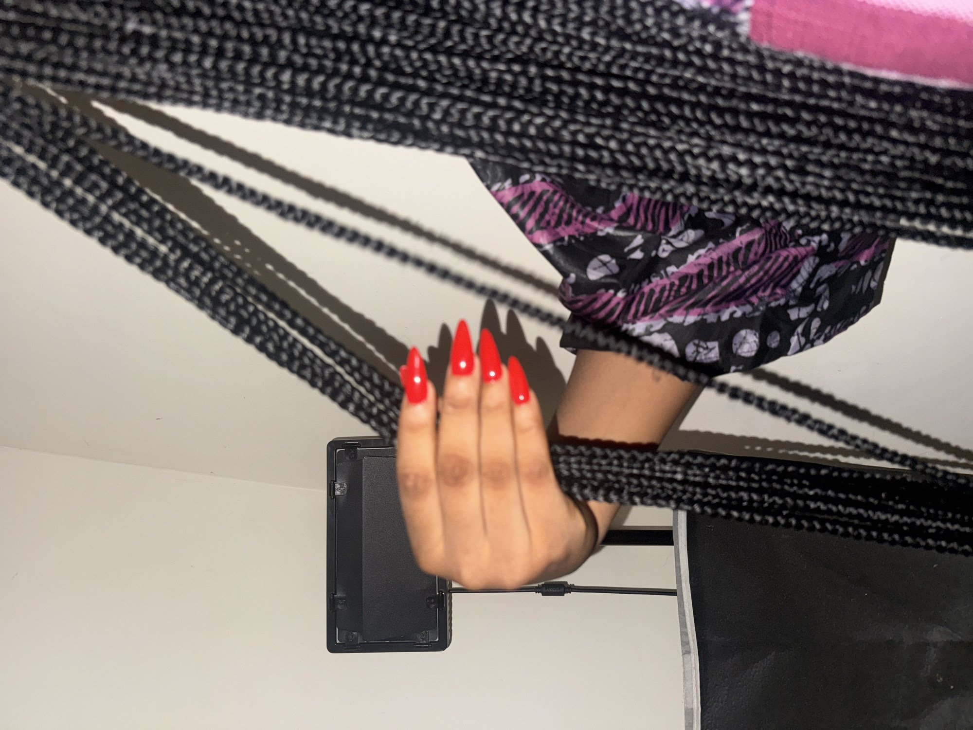 The image presents a close-up view of a person's hand and forearm, set against a plain, light-colored background, likely a wall or flat surface. The primary subject is a hand with long, bright red stiletto-shaped nails, holding a cluster of numerous dark, tightly braided hair extensions. These braids, appearing to be box braids or similar, descend across the frame, casting prominent, elongated shadows on the light background.
A part of the person's clothing is visible on the upper right, featuring a black fabric with an intricate pattern of purple/pink abstract shapes and white detailing. To the left of the hand, a black rectangular electronic device, possibly a power bank or an adapter, rests on the light surface, connected by a thin black cable. Another larger, dark, rectangular object or surface is partially visible in the bottom right corner.
The lighting in the scene appears to be artificial, creating strong contrasts and defined shadows, enhancing the texture of the braids. The overall setting is implied to be indoors, within the location context of Uyo, Nigeria. There is no visible text in the image.