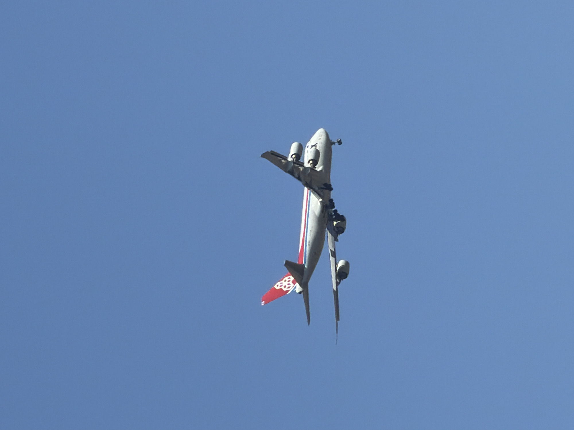 A cargo airplane, specifically a Boeing 747-400BCF, is captured mid-flight against a clear blue sky. The aircraft's tail is prominently displayed, featuring a red and white livery with a distinctive geometric logo. The plane appears to be on approach, as indicated by the deployed landing gear. The image focuses on the aircraft, with no other objects or people visible in the frame. The scene is devoid of any discernible urban or geographical landmarks, making it impossible to identify a specific location within Leudelange, Luxembourg.