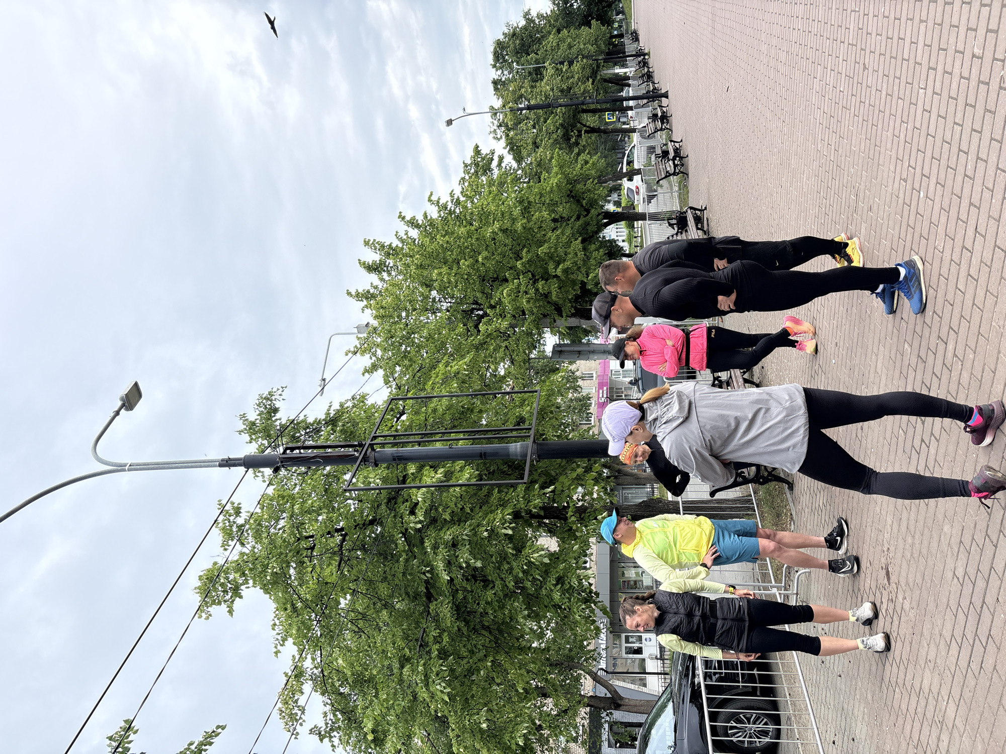 Five people are lined up on a brick sidewalk, appearing to be stretching or exercising. The first two individuals on the right are men, dressed in black athletic wear. The third person is a woman wearing a pink jacket. The fourth person is wearing a grey jacket and a white hat. The last person, to the left, is a woman in a black vest and light green shirt. Behind the group, there is a line of trees along the sidewalk and a row of benches. There are power lines going to a lamp post in the scene. In the background, across from the sidewalk, there are buildings.
The location is in the city of Chaikovsky, Russia.