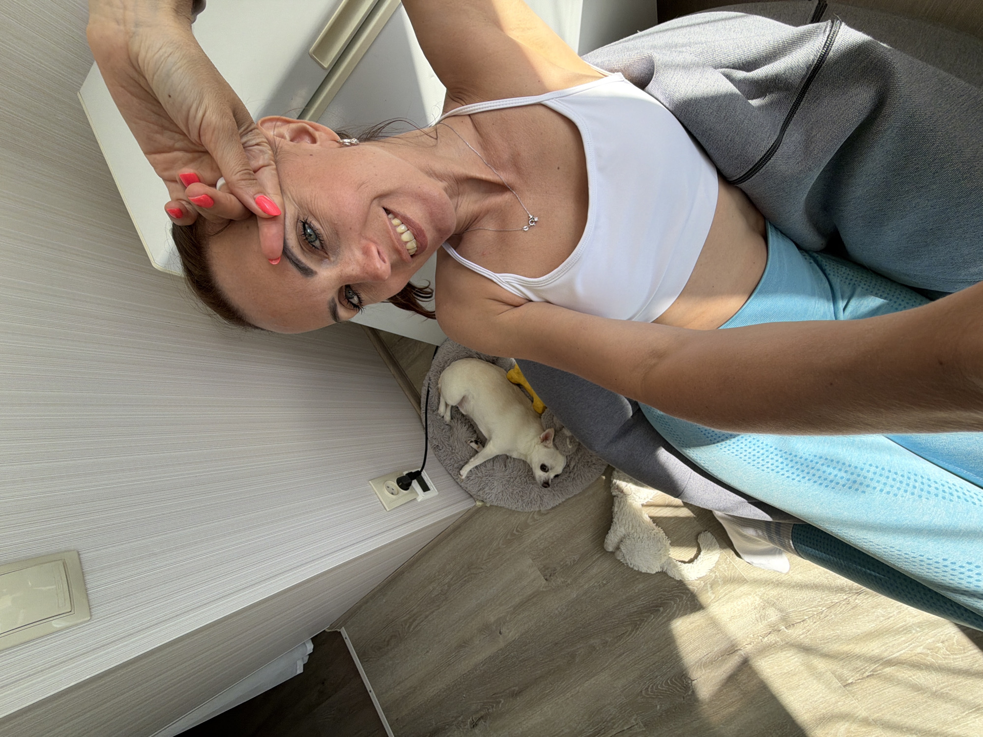 A high-angle selfie shows a cheerful woman smiling broadly, positioned indoors in what appears to be a home setting. She has brown hair, expressive green-blue eyes, and her fingernails are painted a vibrant coral. She is wearing a white sports bra, a delicate silver necklace with a small pendant, and light blue athletic leggings with a subtle dotted pattern. Her left hand is raised, making a peace sign towards her face. A grey jacket or garment is partially visible, draped around her lower body.
To her right, a small, light-colored dog, resembling a Chihuahua, is resting comfortably on a soft, grey pet bed on a light wooden floor. A yellow dog toy lies nearby. An electrical outlet, consistent with European standards (like those found in Russia), is visible on the light grey striped wall next to the dog's bed, with a black cord plugged in. A white fluffy toy or blanket is also on the floor.
Strong, natural sunlight streams into the room, casting clear shadows and illuminating the scene. The overall ambiance is bright and relaxed, suggesting daytime. The woman's wide smile and pose convey happiness and comfort within her home environment, possibly in Birobidzhan, Russia.