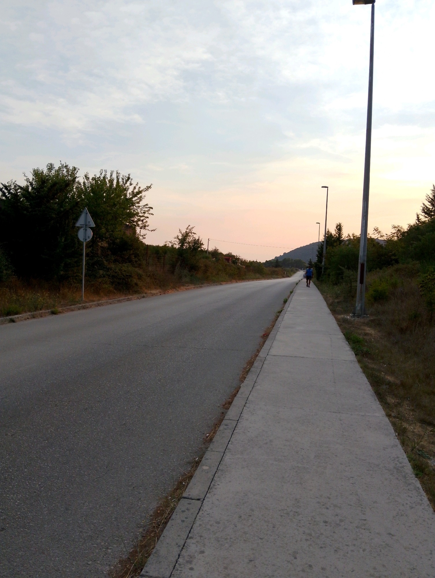 The image shows a paved road and a sidewalk in Sutomore, Montenegro, at dusk. The sky is a pale orange and white, with scattered clouds. On the left side of the road, there is a dense green bush with a traffic sign. On the right side of the road, a sidewalk stretches into the distance, bordered by dry grass and trees. Several streetlights are visible along the sidewalk, with the closest one being a tall, dark pole with a light fixture at the top. In the distance, a person wearing a blue shirt and dark shorts is walking away from the camera on the sidewalk. The road curves gently to the left, leading towards a distant hill covered in trees. The overall scene depicts a quiet, late-day atmosphere in a suburban or rural area of Sutomore.