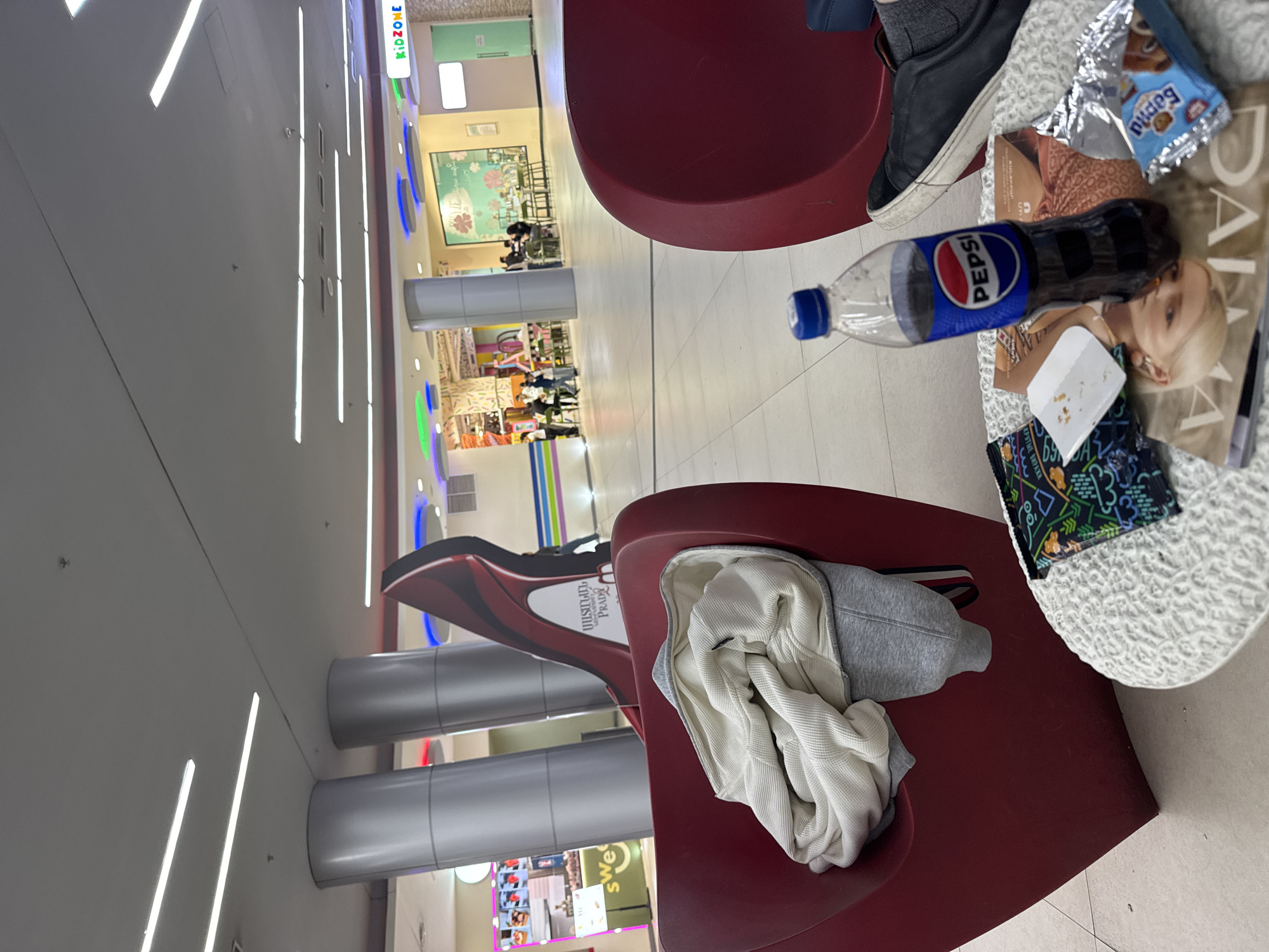 The image depicts a scene inside a shopping mall in Yerevan, Armenia. Several red curved chairs surround a small white table. On the table are a half-empty Pepsi bottle, a magazine featuring a fair-skinned woman with blonde hair, a partially-eaten candy wrapper, and a small packet of food. A pair of dark-colored sneakers rests on the table's edge. One chair has a folded gray and white jacket. In the background, there is a view of various mall shops and activities. People can be seen in the distance, moving through the mall. Overhead are bright fluorescent lights.