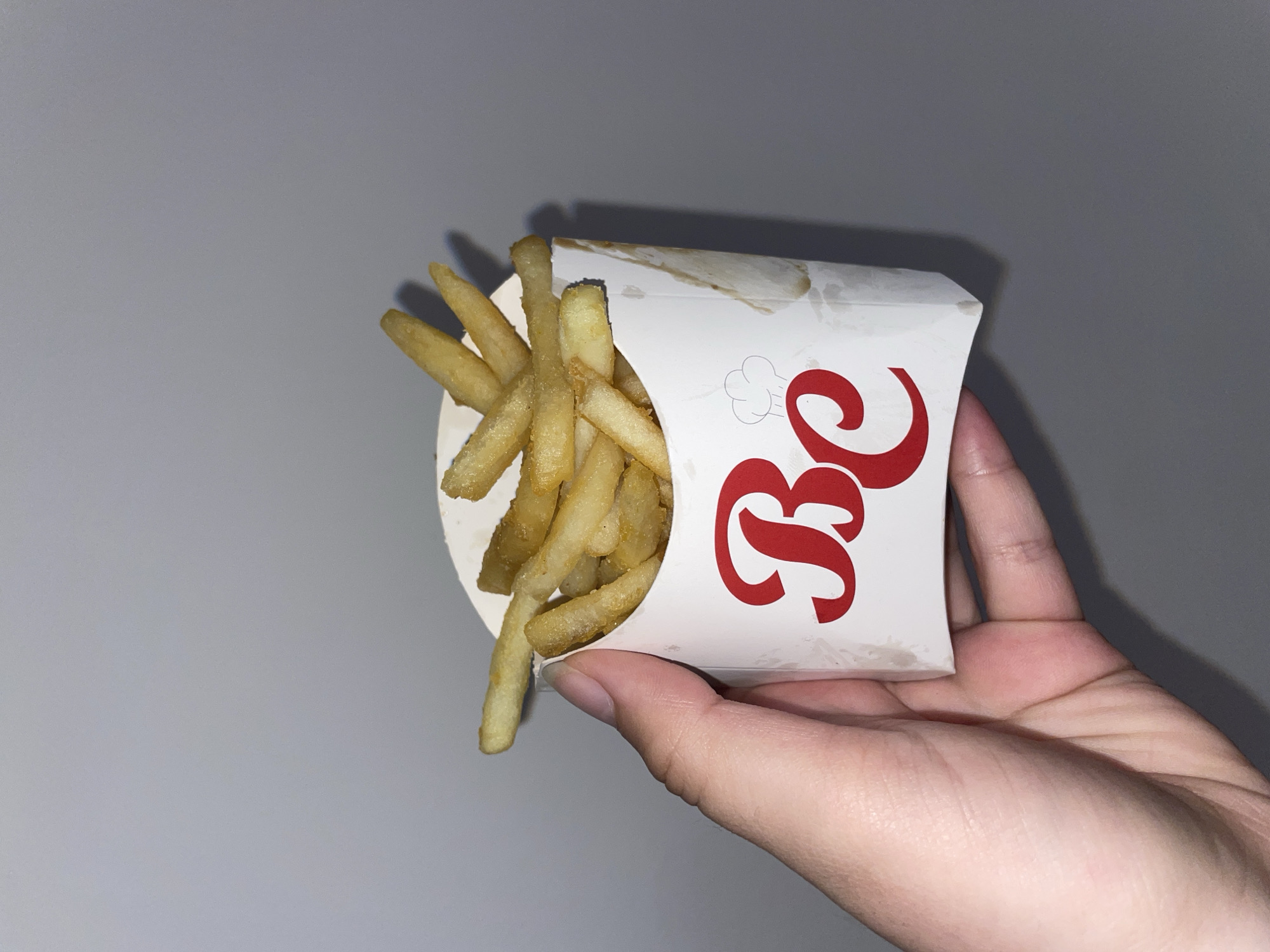A person is holding a white carton of french fries. The carton has a red logo with the letters "Bc" on it. The fries are overflowing from the carton. The background is a plain gray surface. There is no visible information that specifies the location of the city of Plovdiv, Bulgaria.