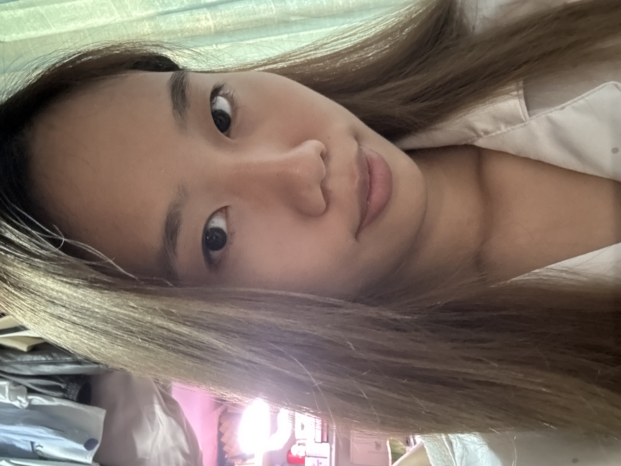 A close-up, slightly tilted selfie of an Asian woman with long, light brown hair is presented. She is wearing a light-colored shirt. Behind her, out of focus, is a glimpse of what appears to be a room with indeterminate objects and a window allowing light to enter. There is no definitive evidence to support the location is in Nan'an District, China.